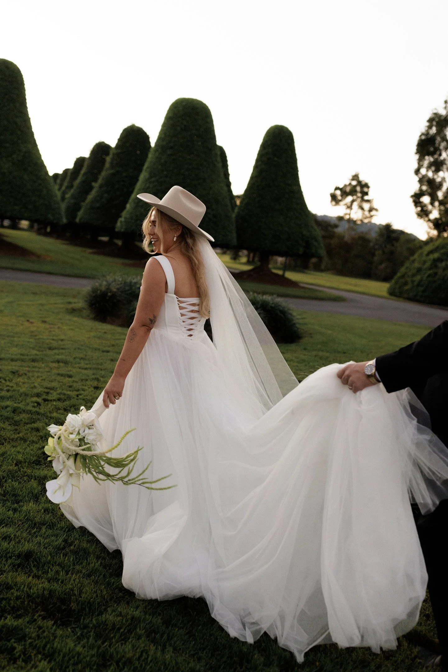A woman in a white wedding dress and a wide-brimmed hat is smiling and holding a bouquet, standing outdoors on green grass with neatly trimmed topiary trees in the background.