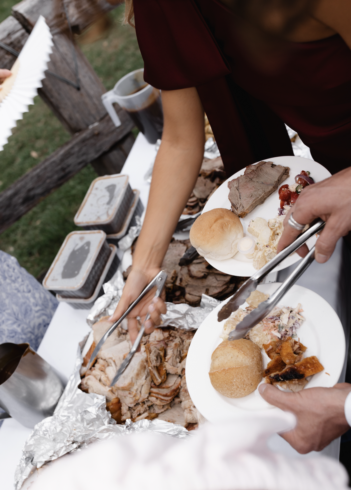People serving and eating barbecue with plates containing sliced meat, rolls, and side dishes at an outdoor gathering.