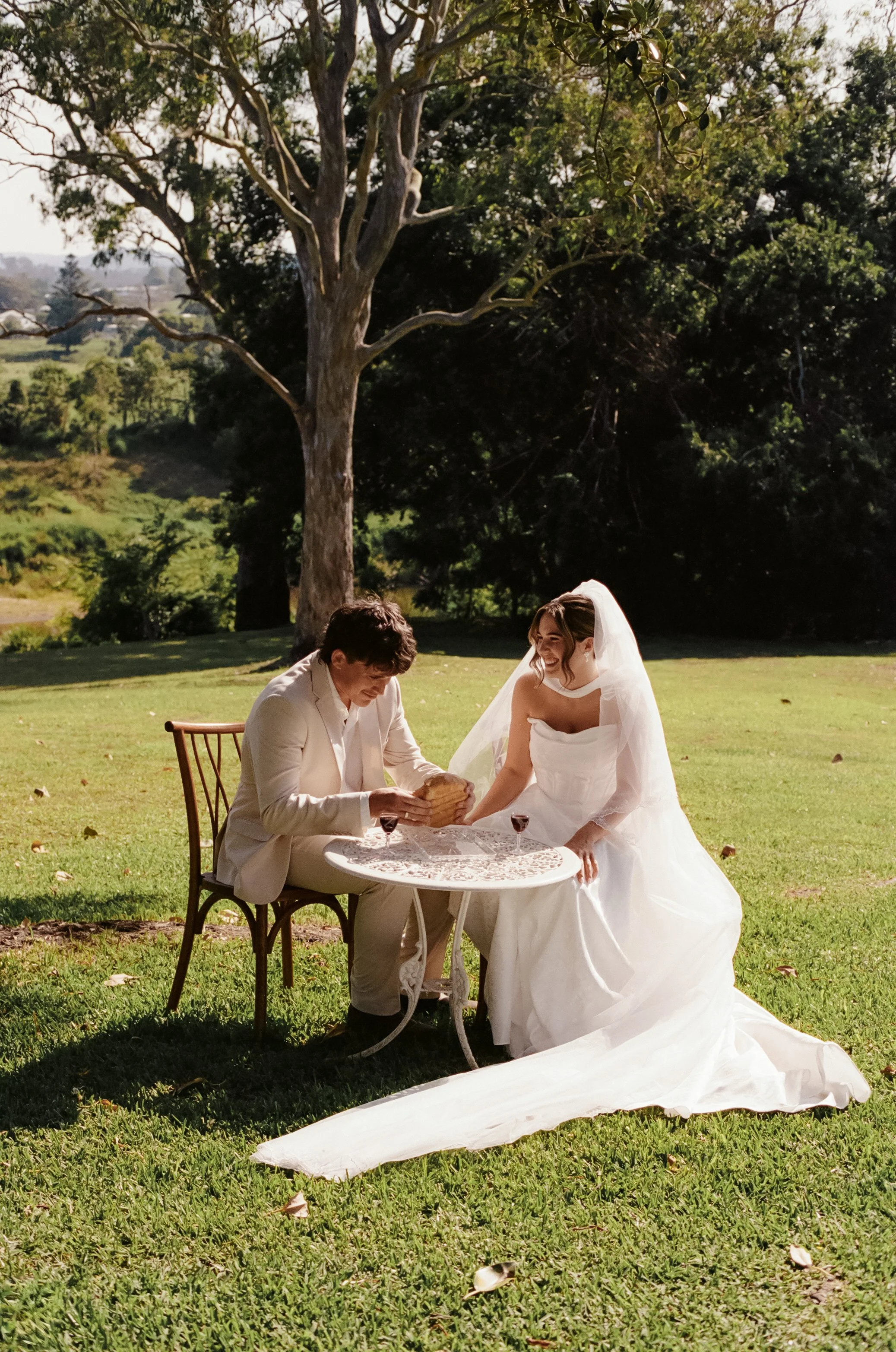 A bride and groom sit at a small round table outdoors on a grassy lawn, sharing a moment with the bride smiling and wearing a wedding dress and veil, while the groom is pouring a drink, faced towards her, with a large tree and greenery in the backgro