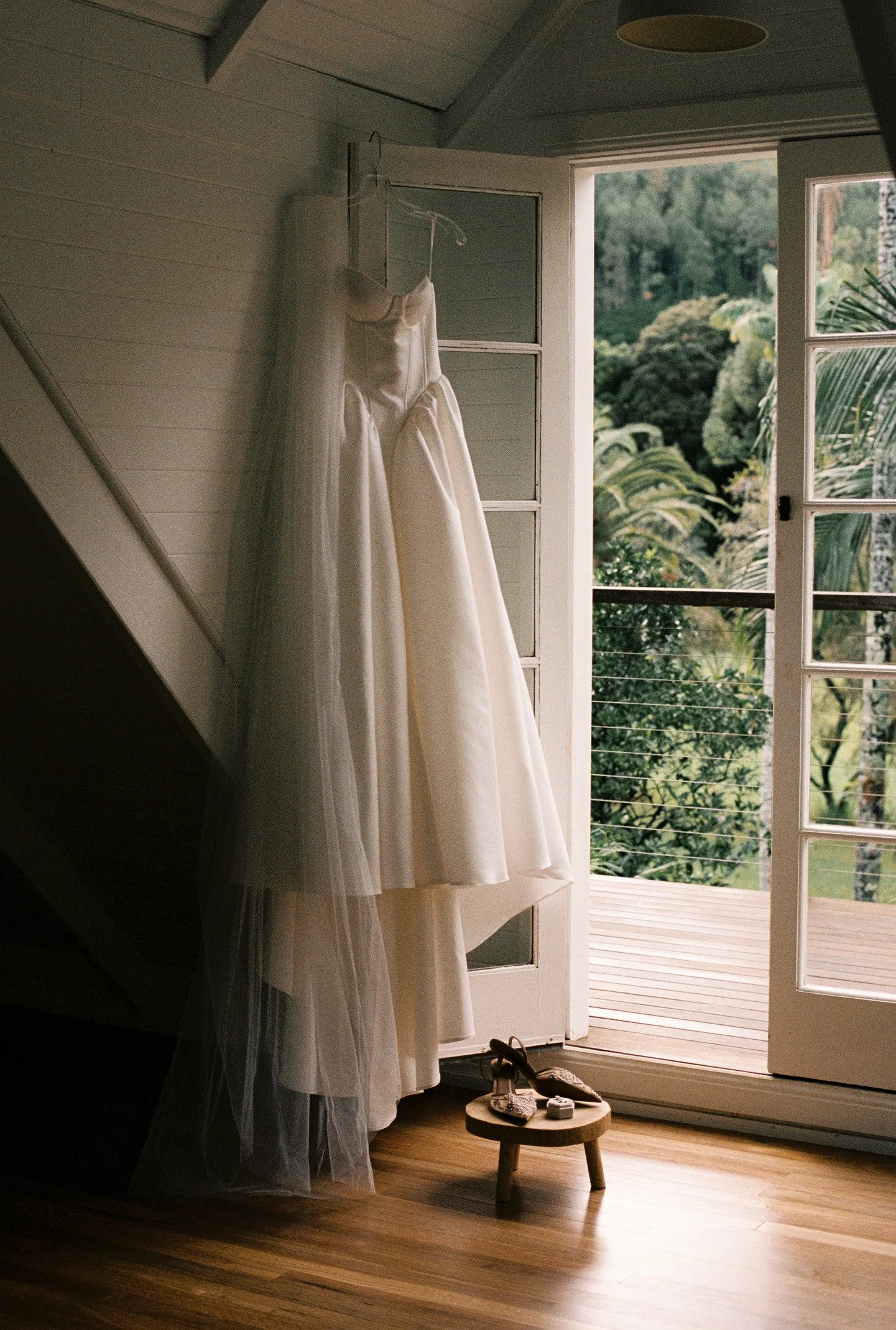 A wedding dress hanging by the open door of a room, with sunlight coming in from outside onto a small stool with bridal shoes and accessories. The outdoors is lush with greenery.