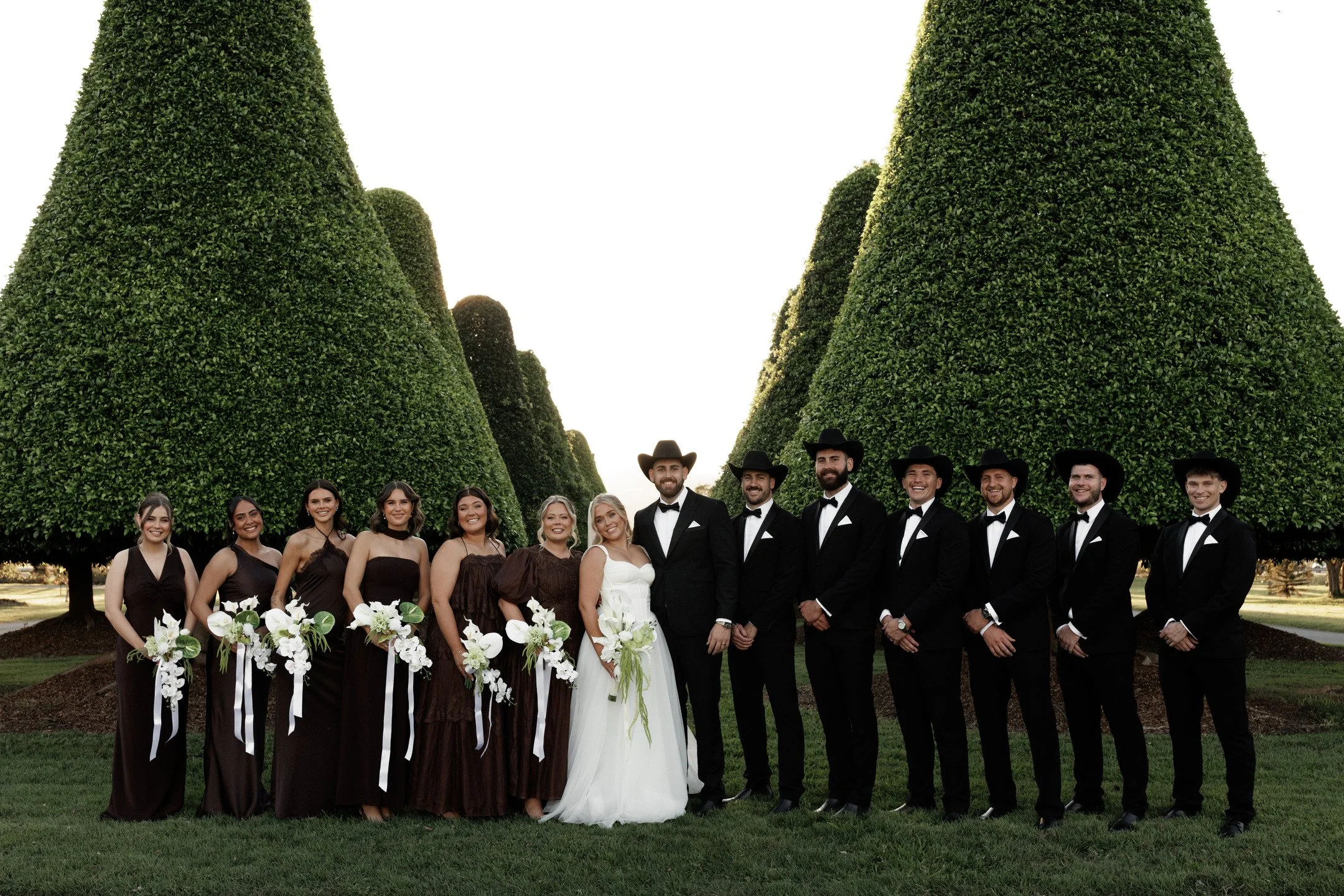 A group of wedding attendants, including bridesmaids and groomsmen, standing outdoors in front of neatly trimmed large green trees, dressed in formal wedding attire, smiling for the photo.