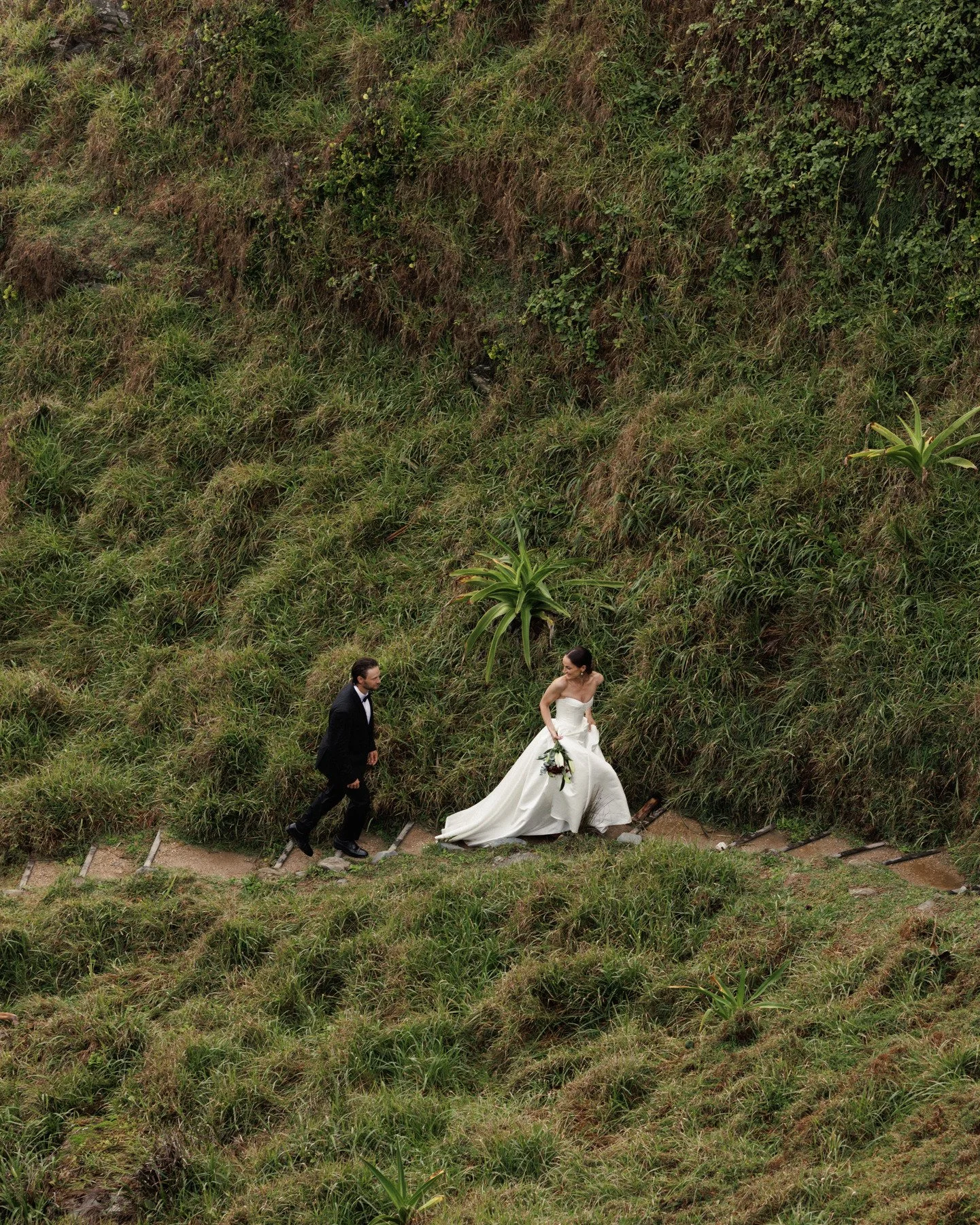 A bride in a white wedding dress and a groom in a tuxedo walk together on a stone pathway in front of a grassy hill with ornamental plants. The bride holds a bouquet and smiling at the groom, while he looks at her.