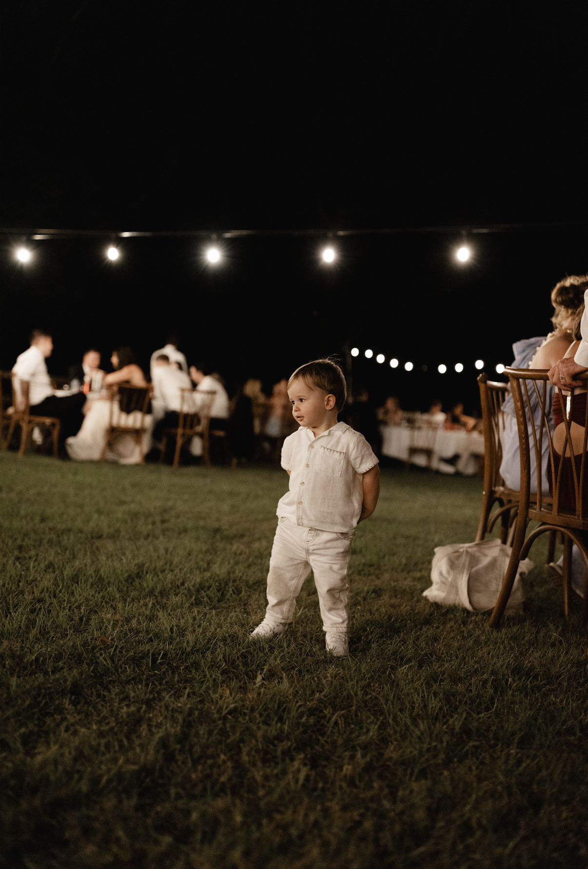 A young boy in light-colored clothing stands on a grassy area at night with string lights overhead and a blurred background of people sitting at tables.