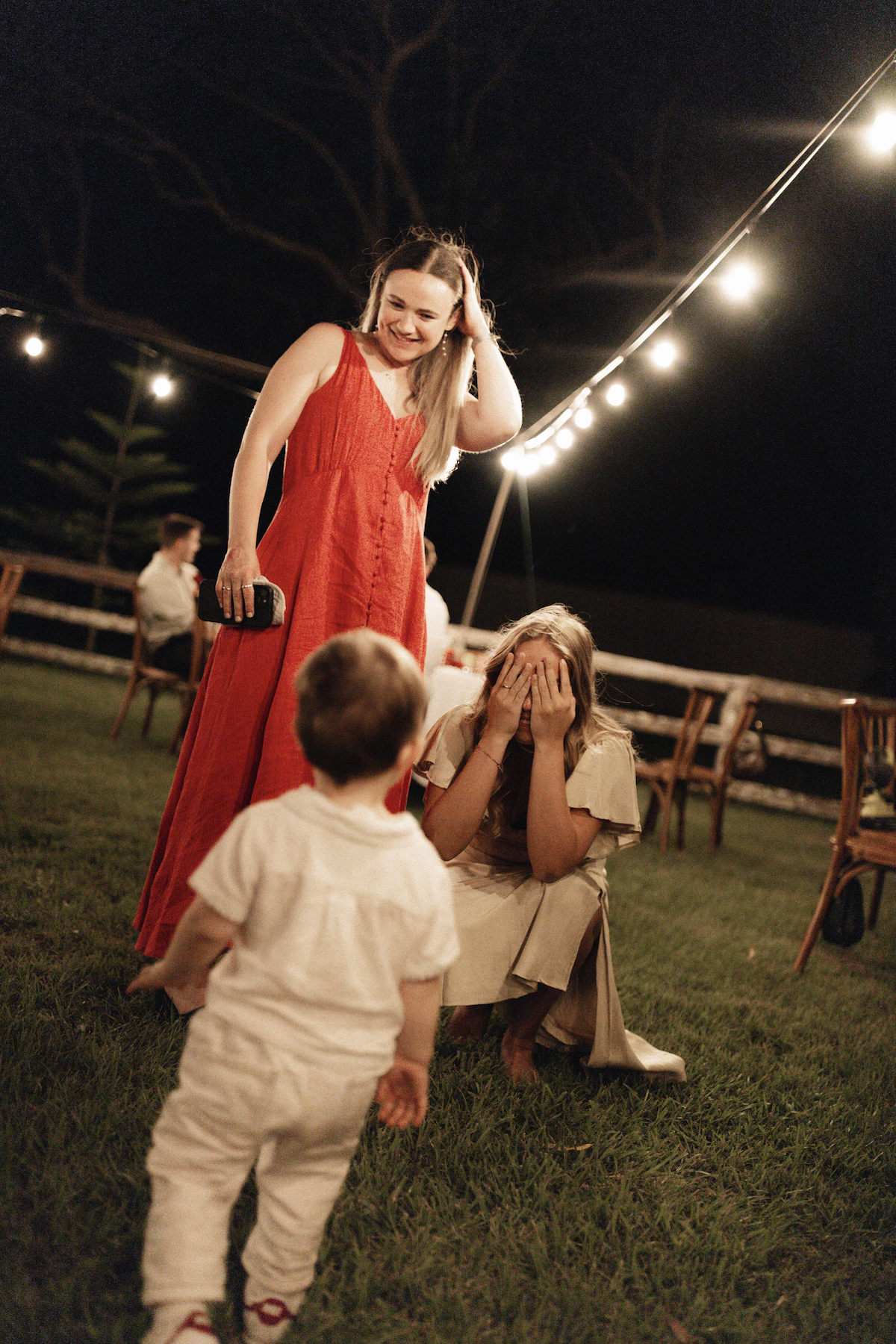 A woman in a red dress smiling and touching her hair stands next to a girl sitting on the ground covering her face with her hands. A young boy stands in front of the girl, facing her. They are outdoors at night under string lights with wooden chairs 