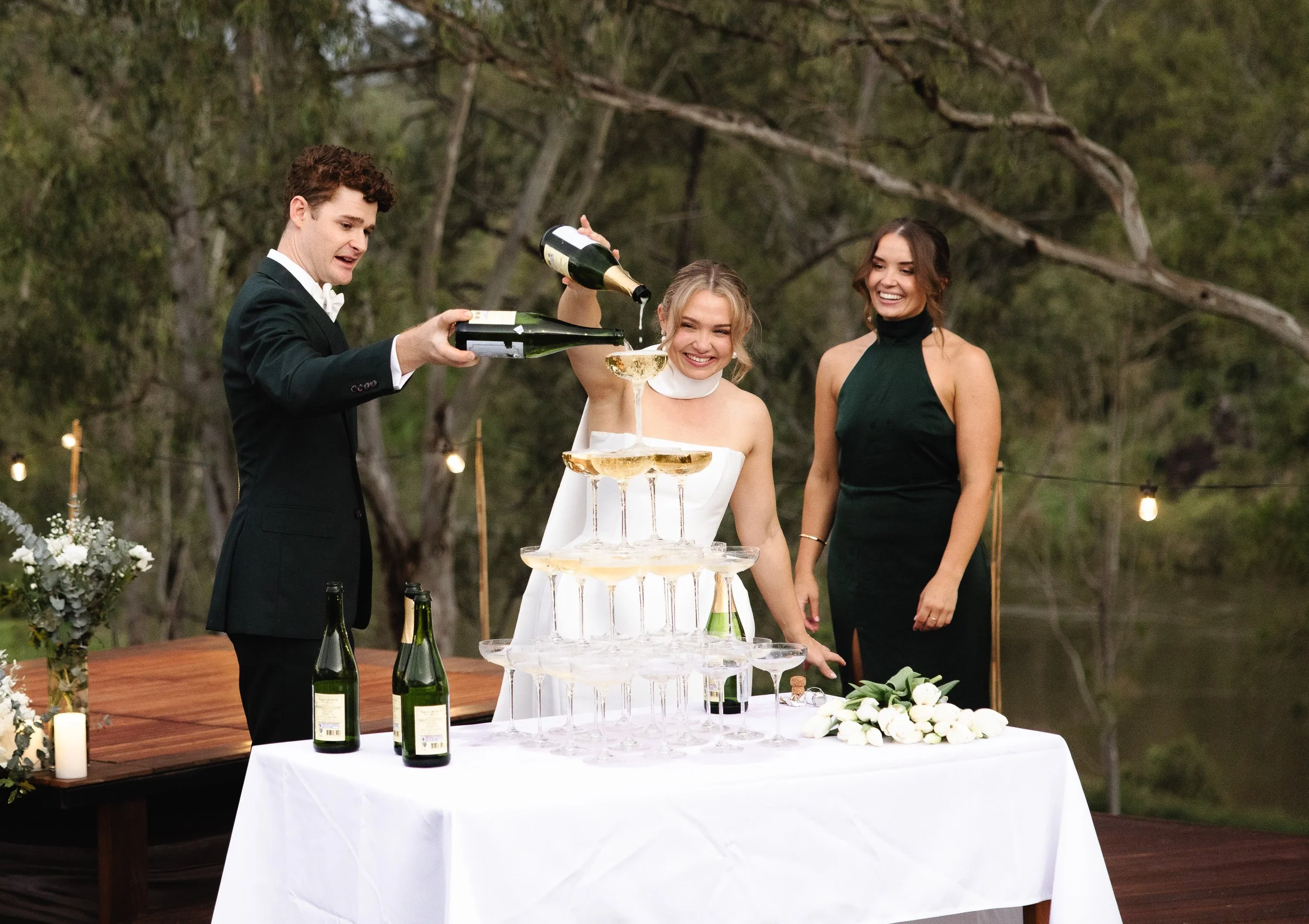 Three people at a wedding pouring champagne into a champagne tower outdoors -Elevated Weddings