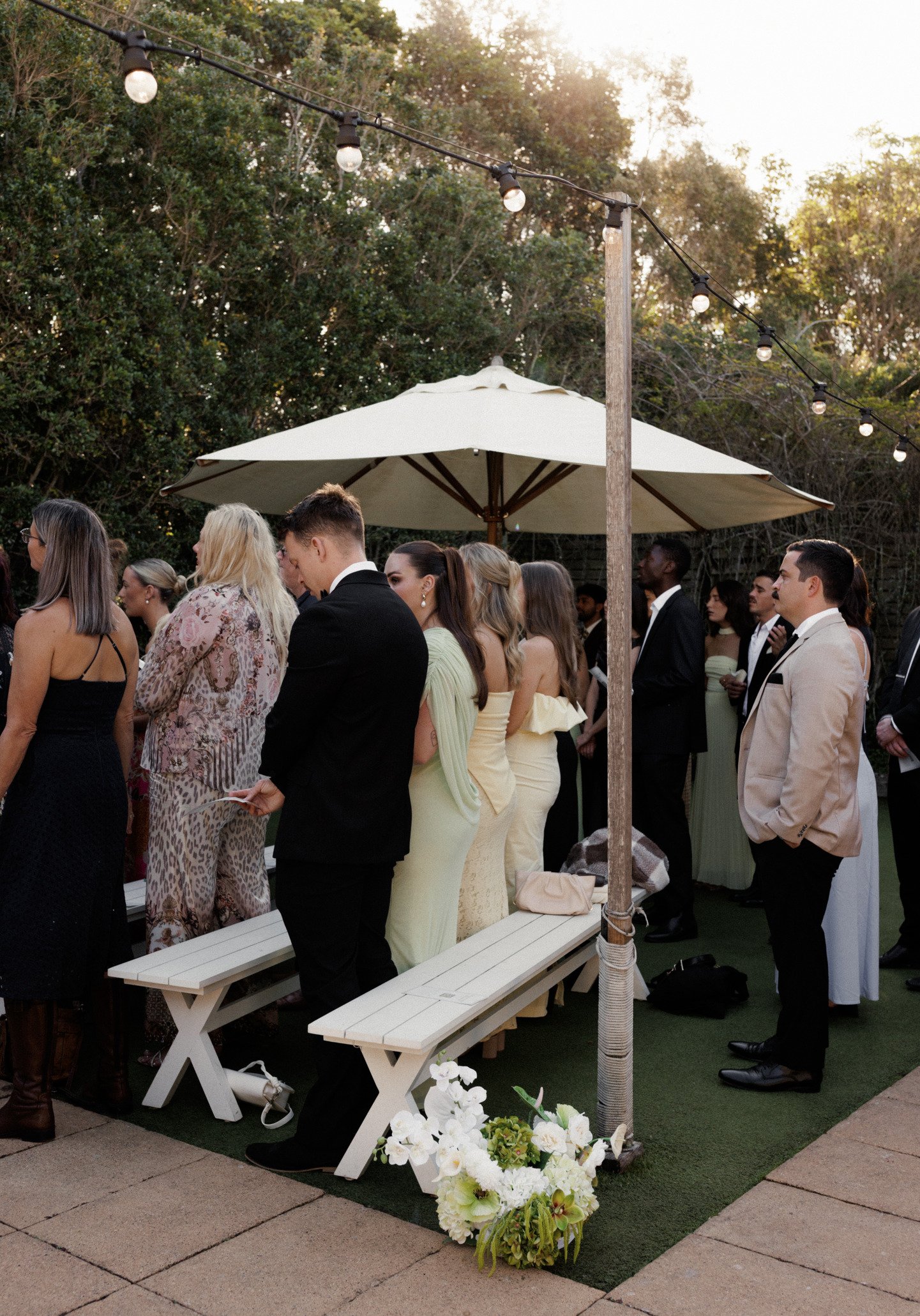 People dressed in formal attire attending an outdoor wedding ceremony under a large umbrella with string lights and flower arrangements.