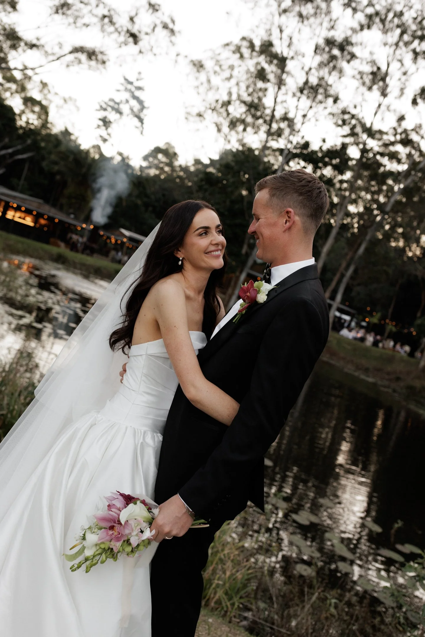 A bride and groom are smiling and looking at each other, standing outdoors near a water body with trees in the background, during sunset or twilight.
