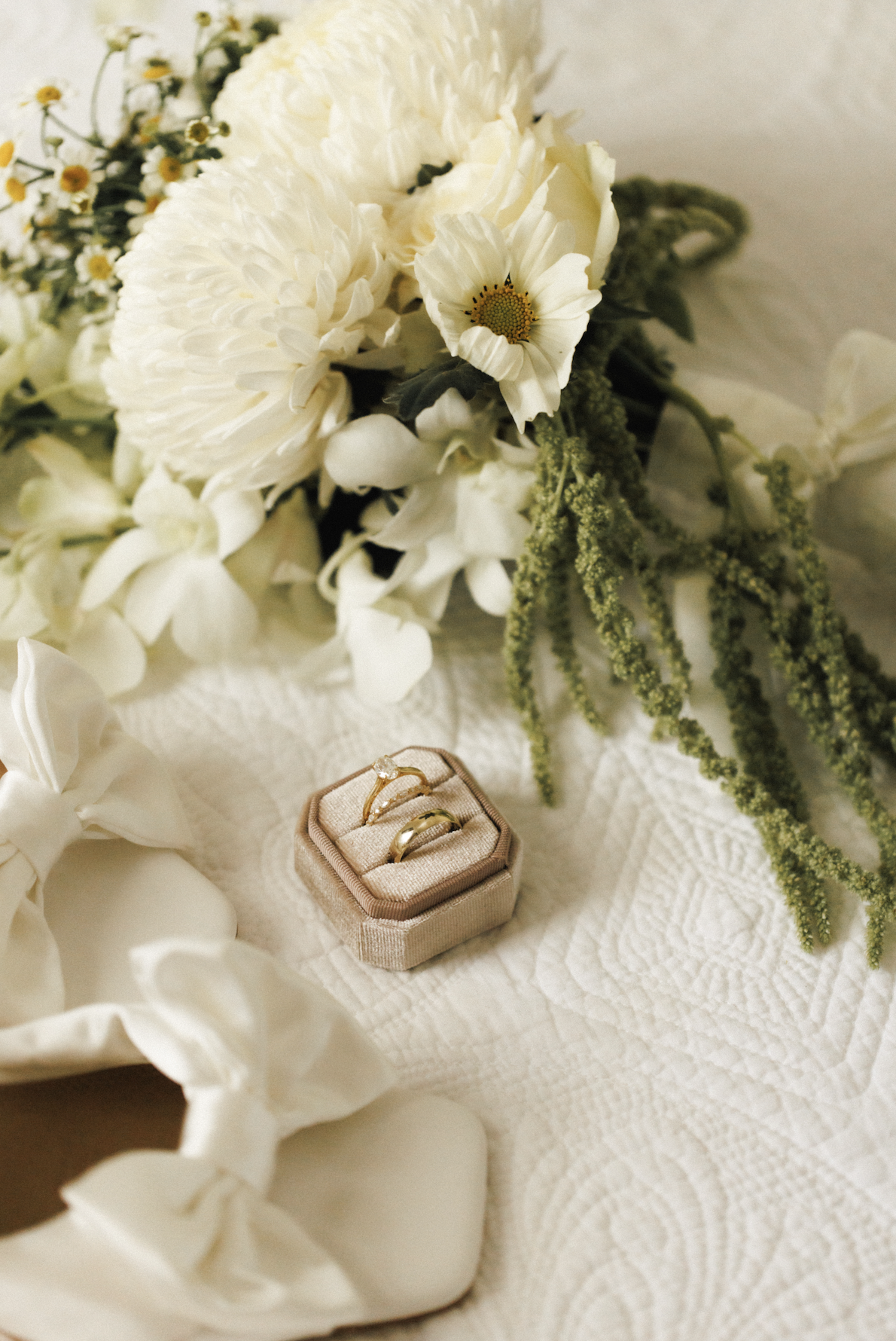 White wedding bouquet with white and green flowers, a beige ring box with two gold rings, placed on a white textured cloth.