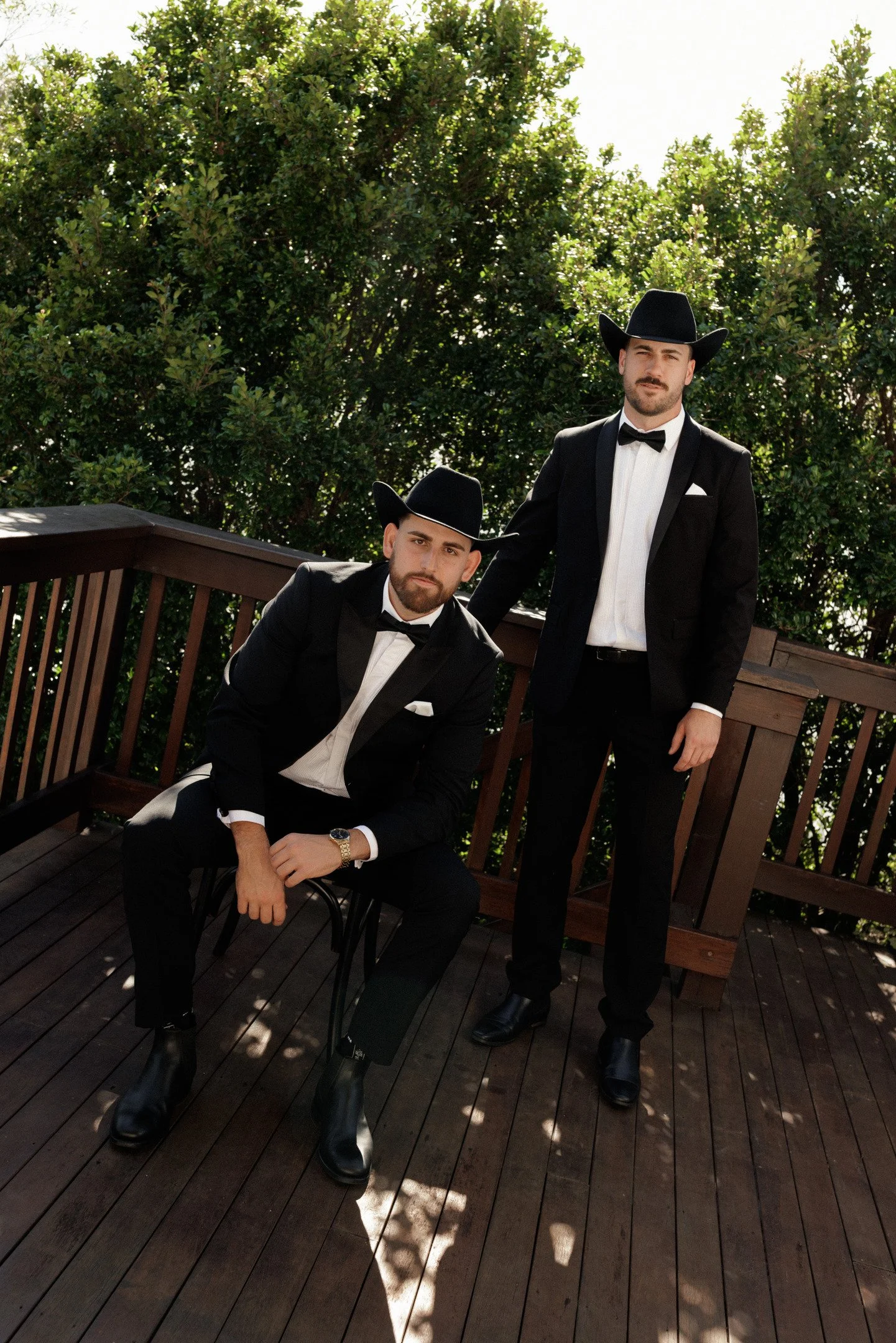 Two men dressed in black tuxedos, white shirts, black bow ties, and cowboy hats, posing on a wooden deck with green trees in the background. One man is sitting while the other is standing.