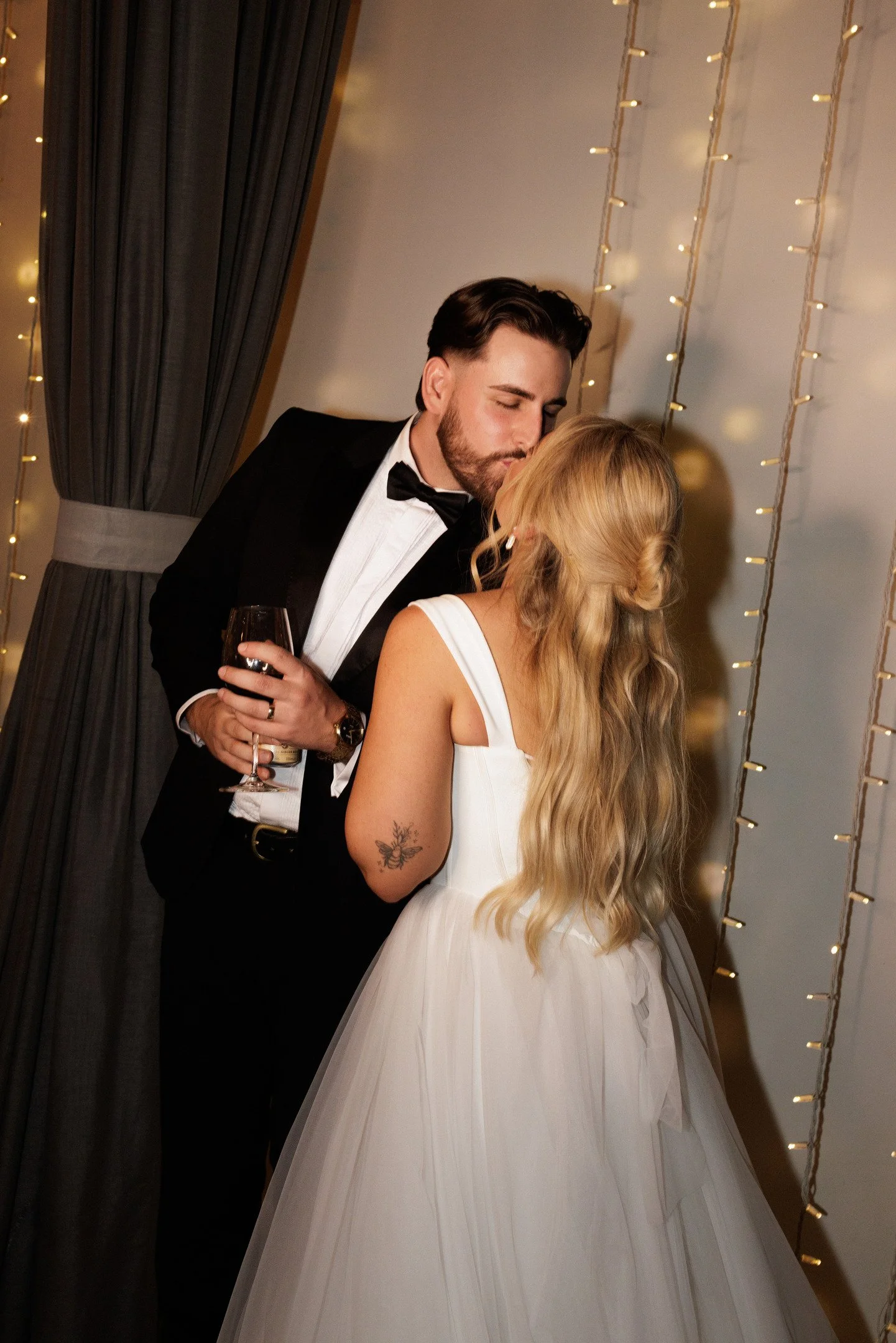 A man in a tuxedo kissing a woman in a white gown at a wedding reception with string lights in the background.