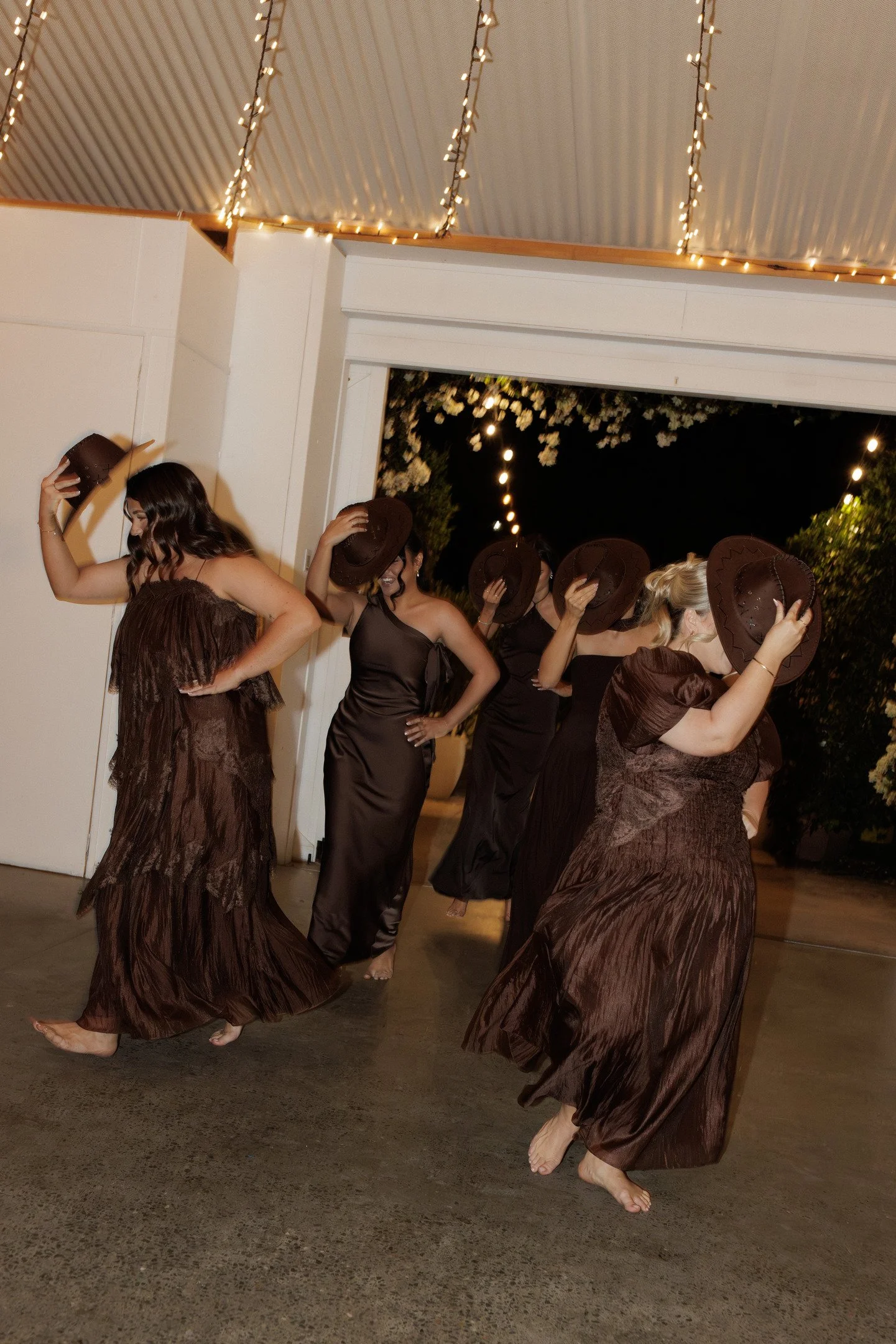 A group of women in dark brown dresses and cowboy hats participate in a dance indoors at night, with string lights hanging from the ceiling and trees visible through an open door.