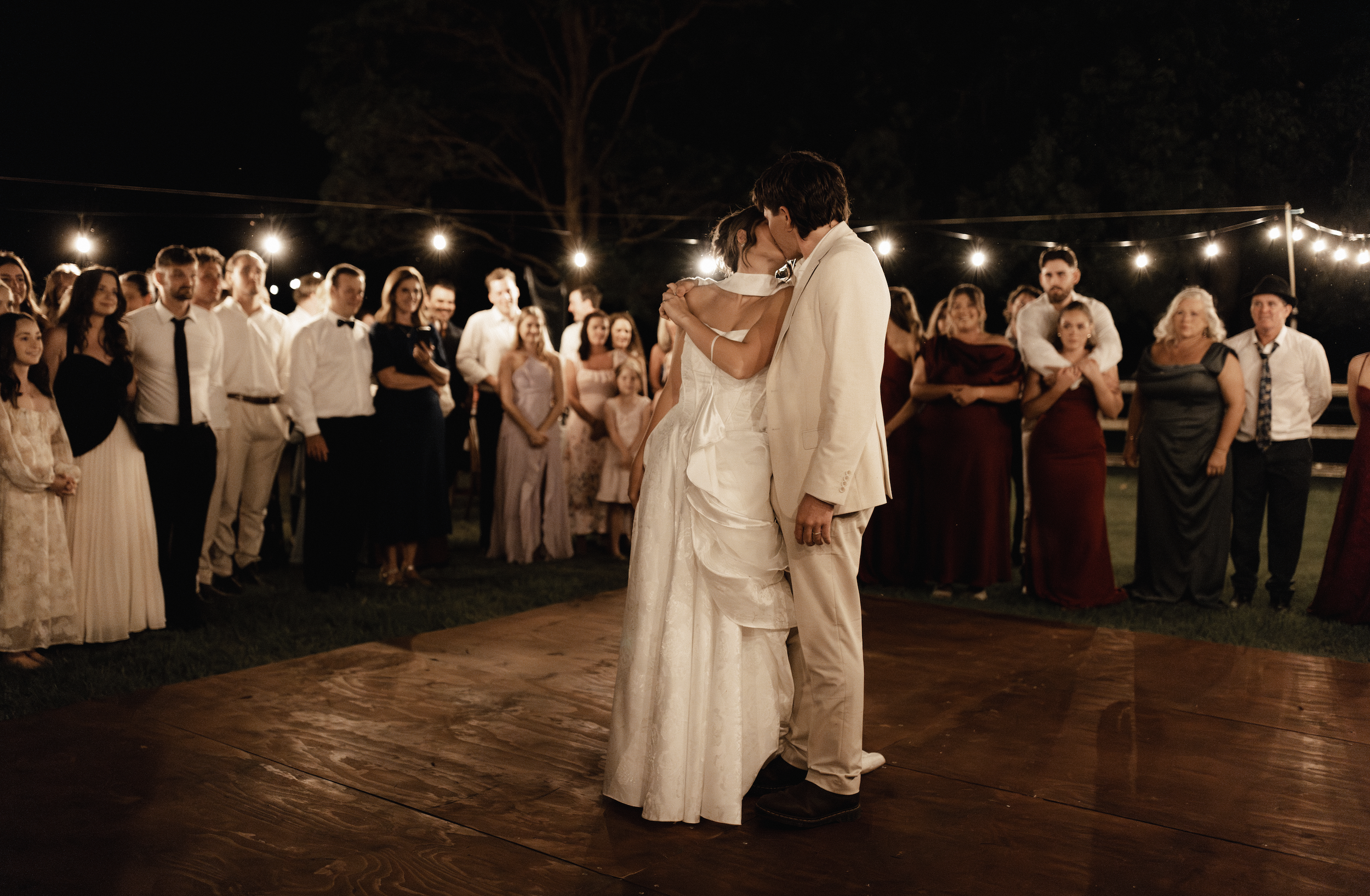A couple shares a first dance at their wedding reception on a wooden dance floor at night, surrounded by friends and family, with string lights overhead.
