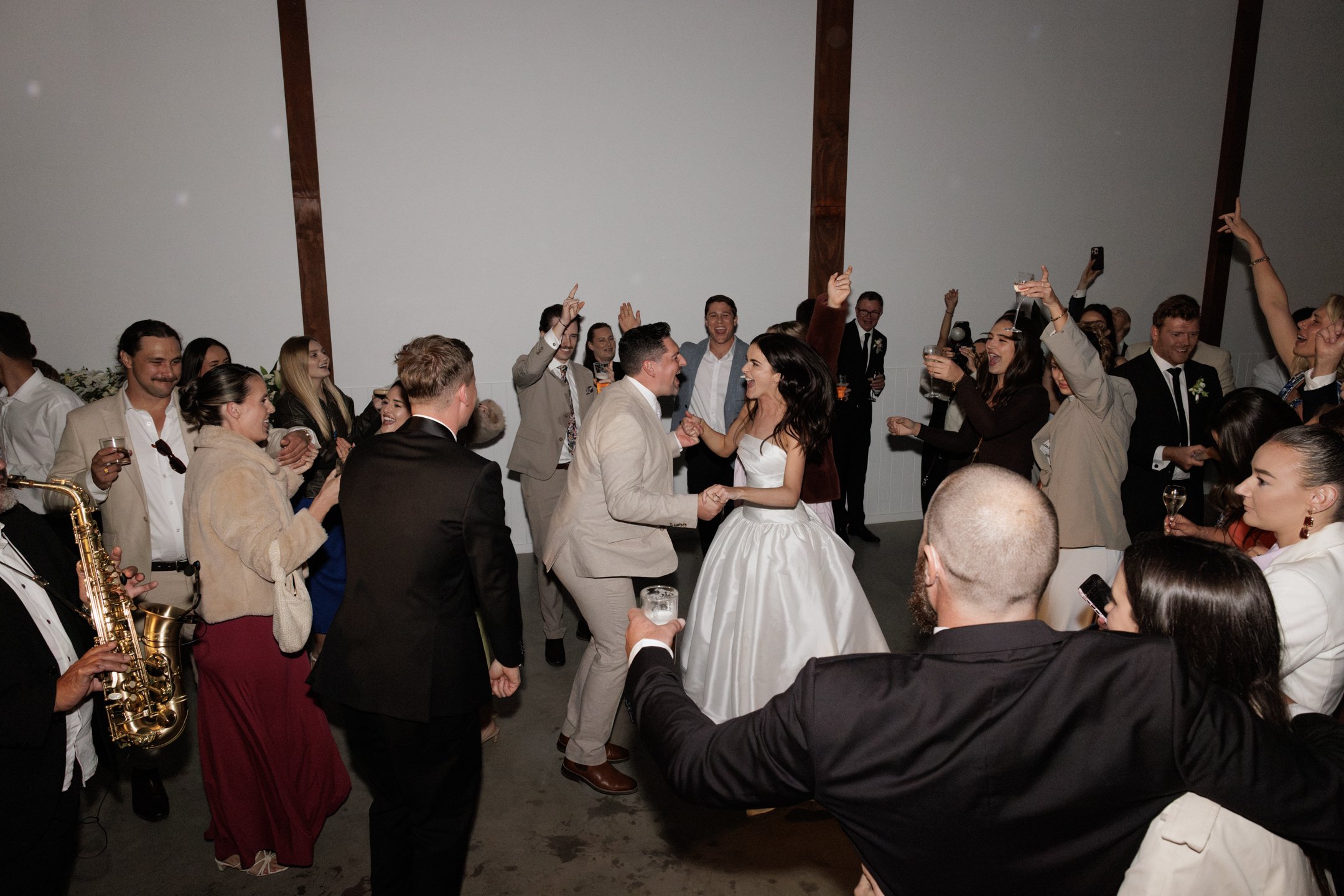 People dancing and celebrating at a wedding reception, with the bride and groom dancing in the center and guests around them enjoying drinks and music.