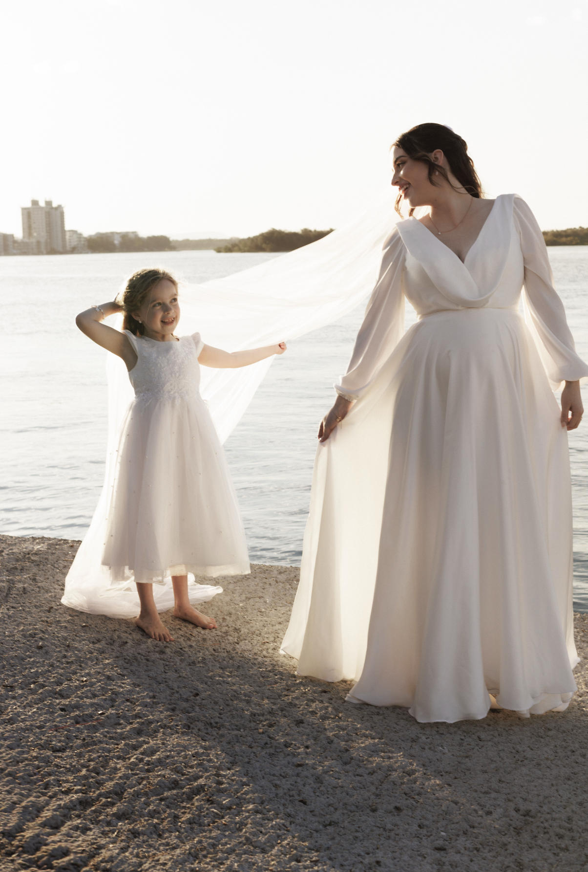 A woman and a girl in white dresses near a body of water at sunset, holding a flowing fabric.