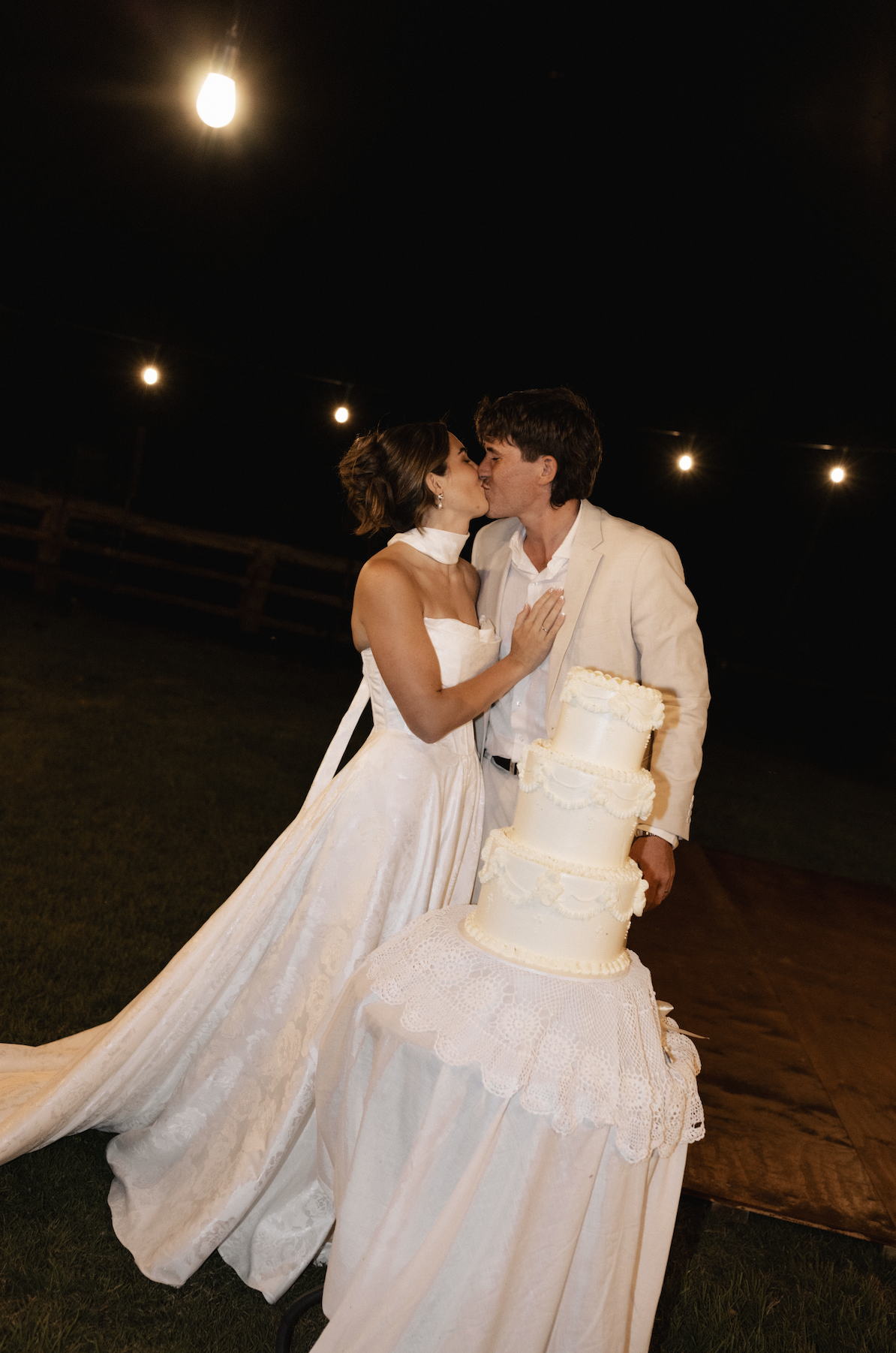 A newlywed couple sharing a kiss at night during their wedding celebration, standing next to a three-tiered wedding cake on a decorated table with a lace cloth, outdoor setting, string lights overhead.
