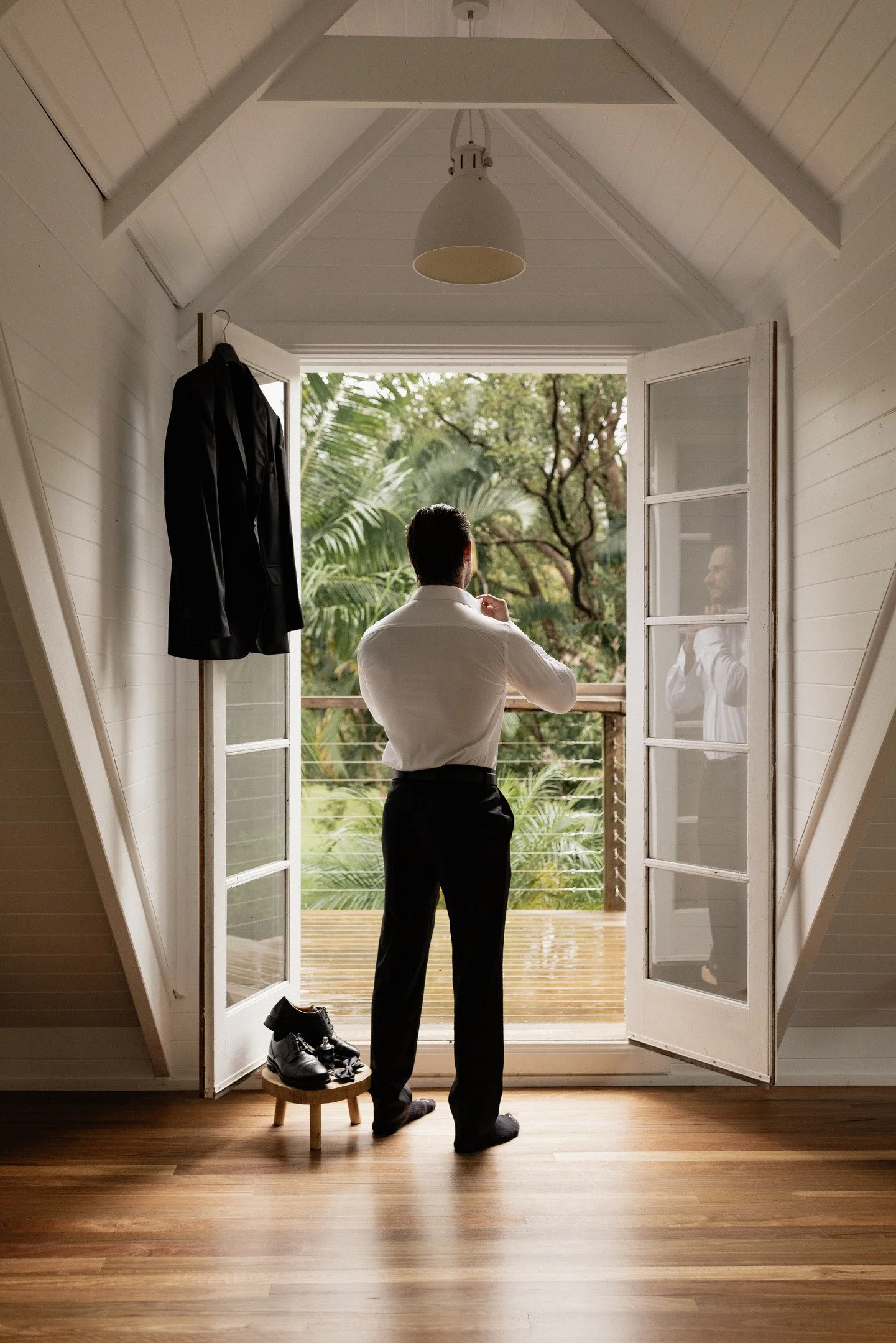 A man dressed in a white shirt and black pants stands on a wooden floor inside a small, white, wooden room with an open door, looking outside at swaying trees and a lush, green garden. On a small round stool nearby, there are black dress shoes and a 