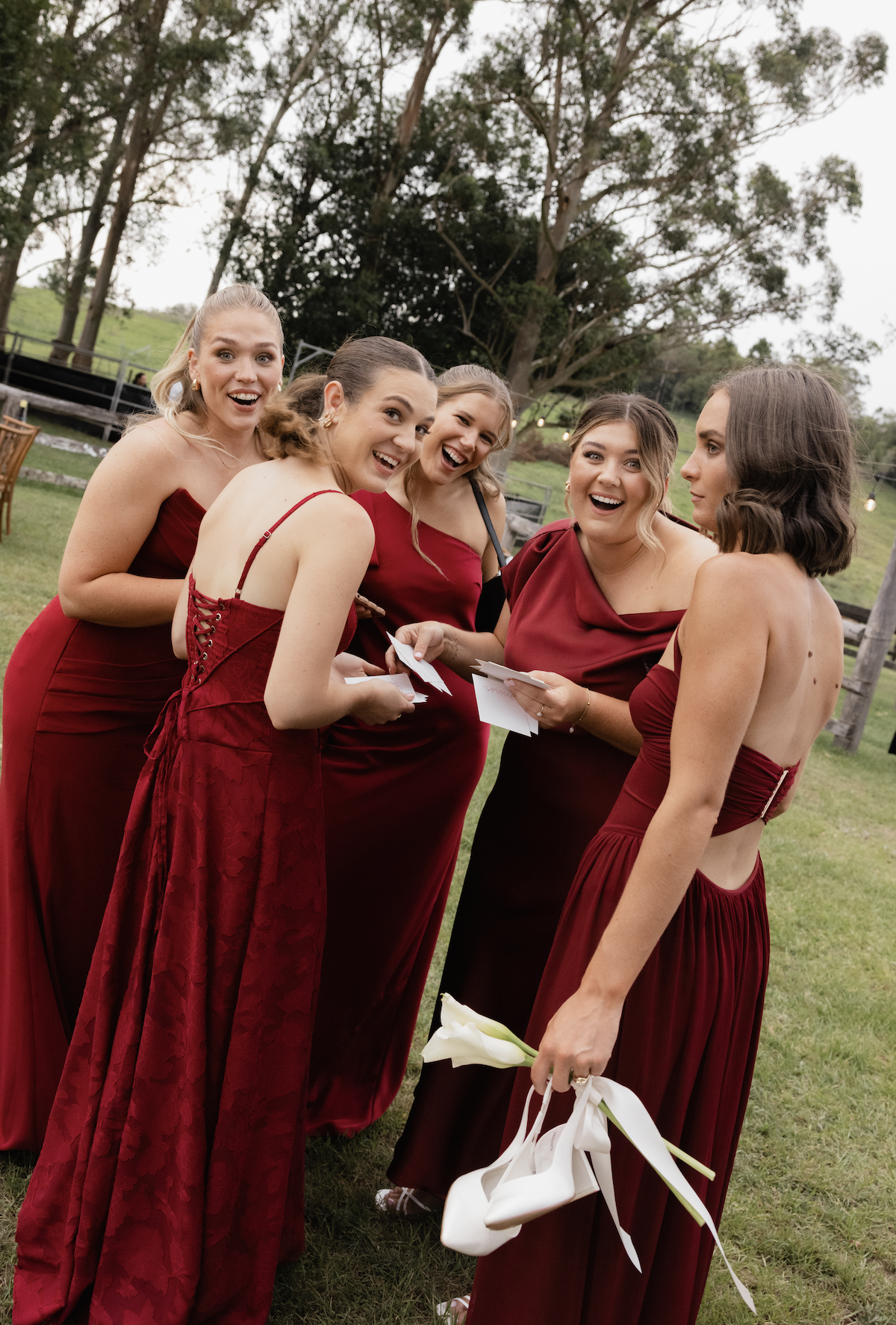 Five women in red dresses laughing and talking outdoors at a wedding or celebration, with trees and cloudy sky in the background.