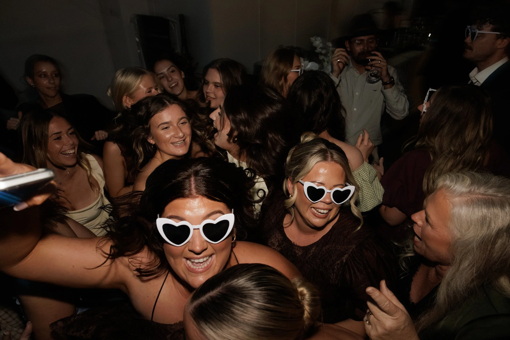 A group of people at a party dancing, smiling, wearing glasses, with a dark background and a lively atmosphere.