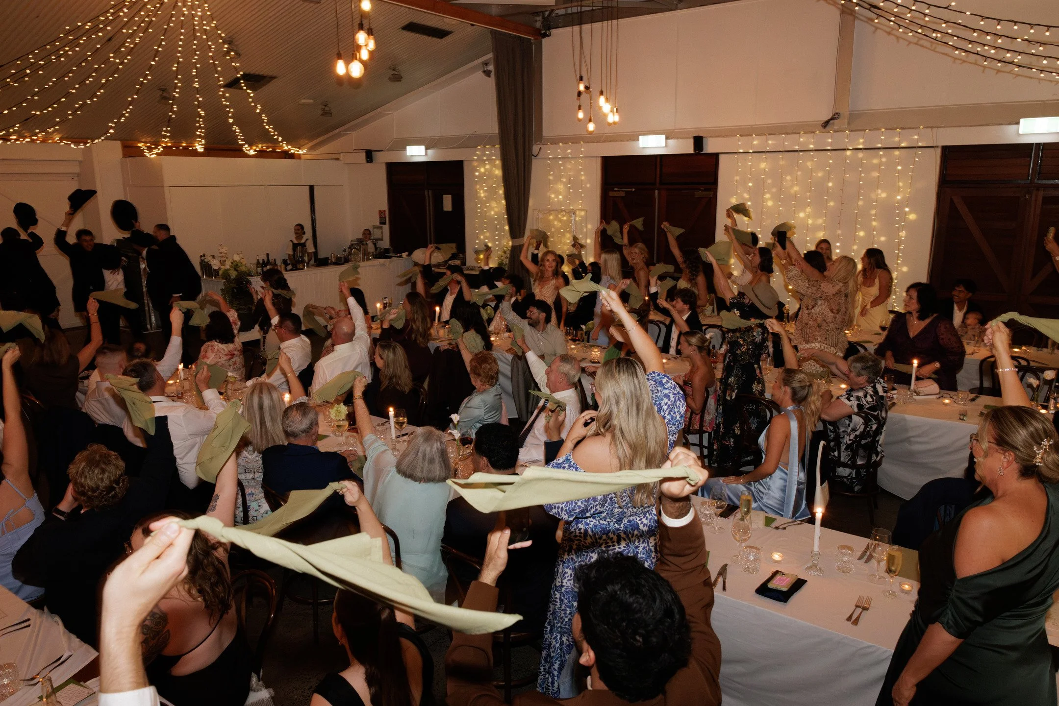 A large indoor wedding reception with many guests seated at decorated tables, some guests are standing and waving napkins, a string of lights decorates the ceiling, and a bar with bartenders is visible in the background.