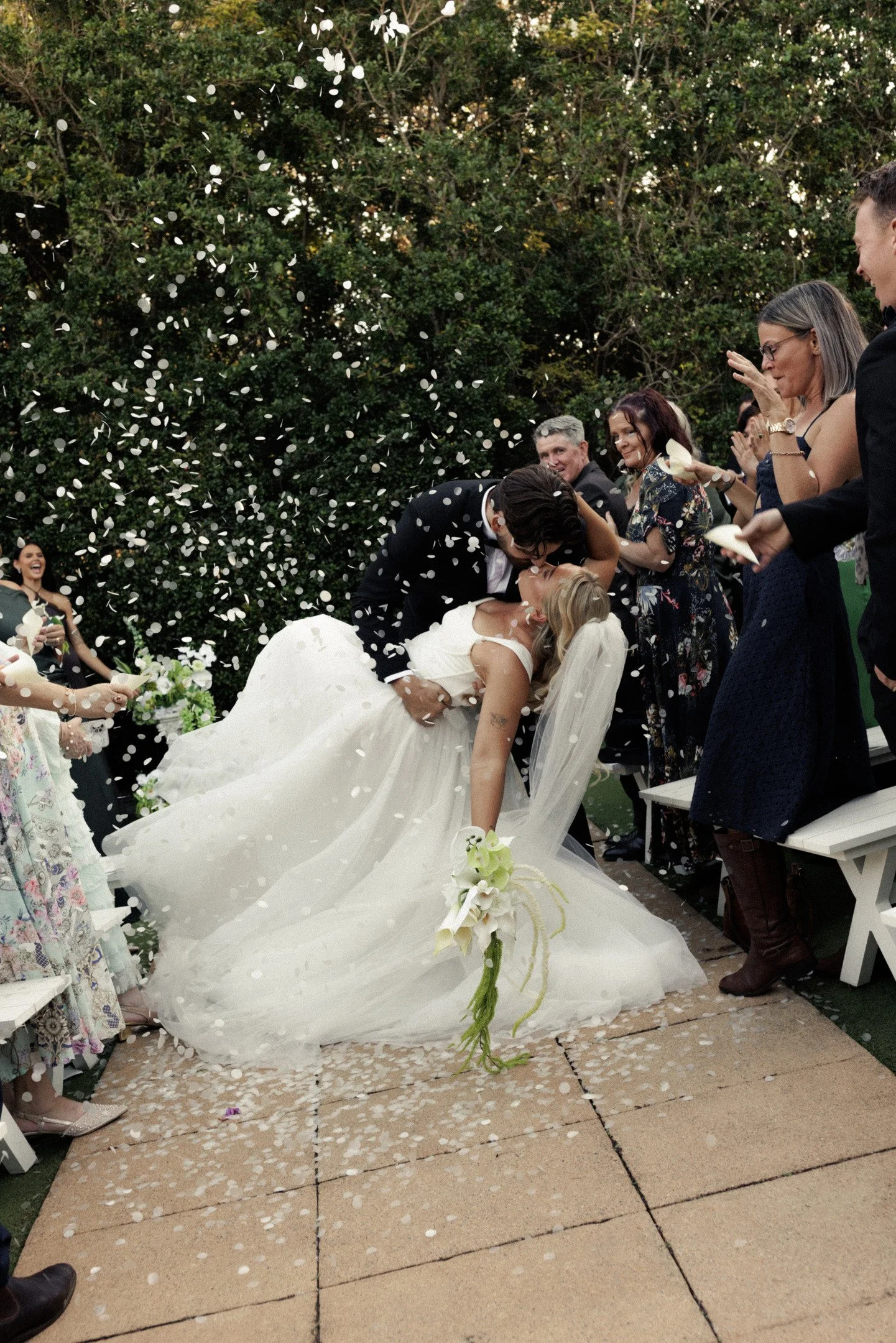A wedding ceremony outdoors with a bride and groom kissing while surrounded by friends and family. White flower petals are falling from above.