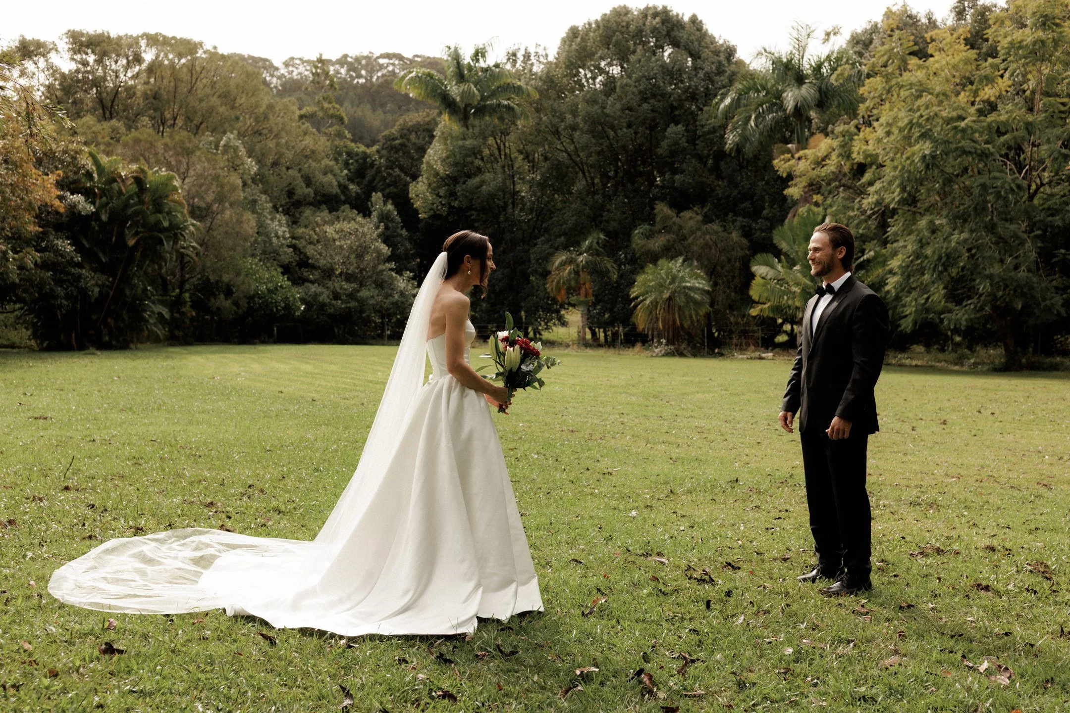 A bride and groom standing on a grassy field facing each other, with the bride holding a bouquet of flowers, surrounded by lush trees.