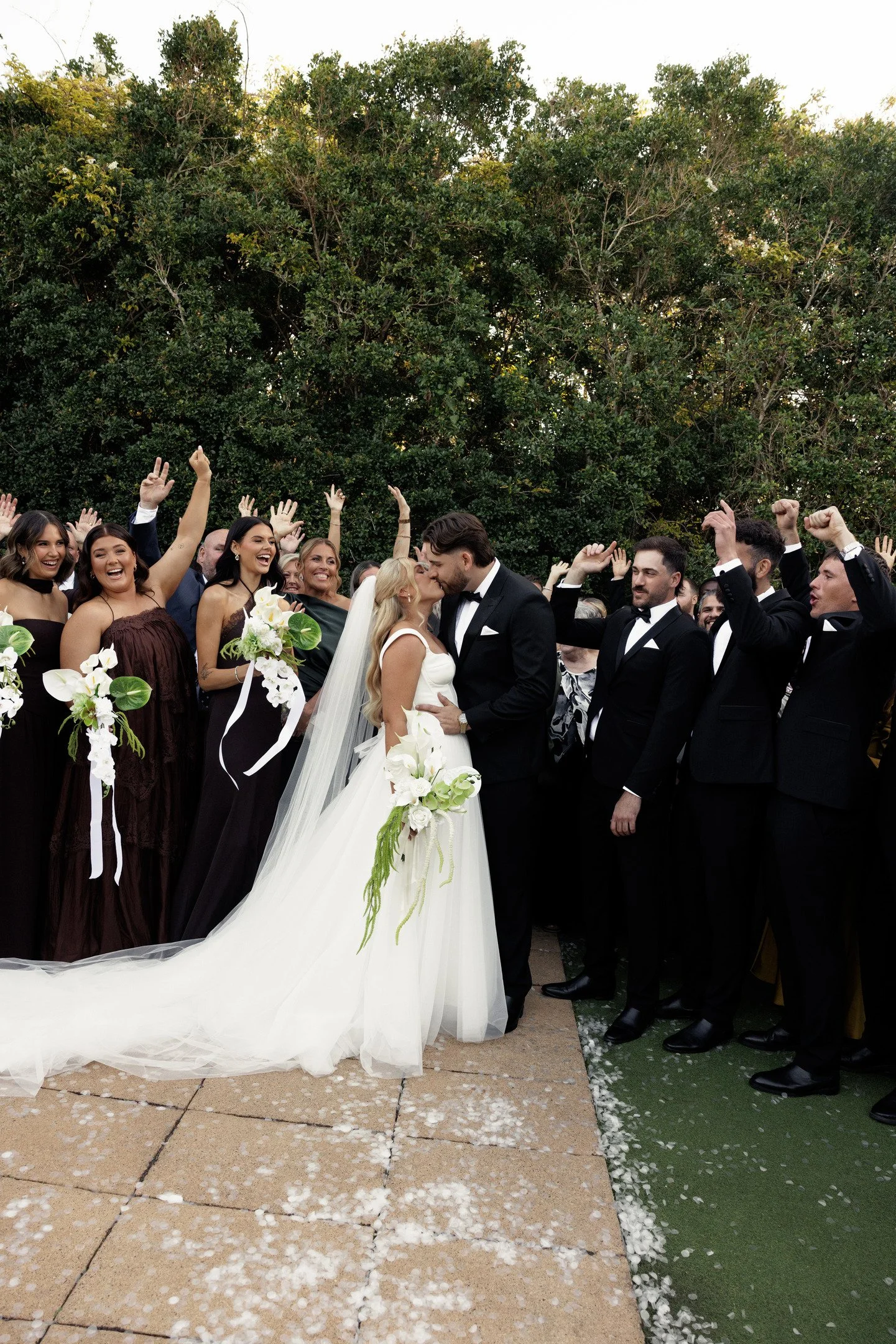 A wedding celebration outdoors with the bride and groom kissing under a green arch, surrounded by friends and family celebrating and throwing flower petals.