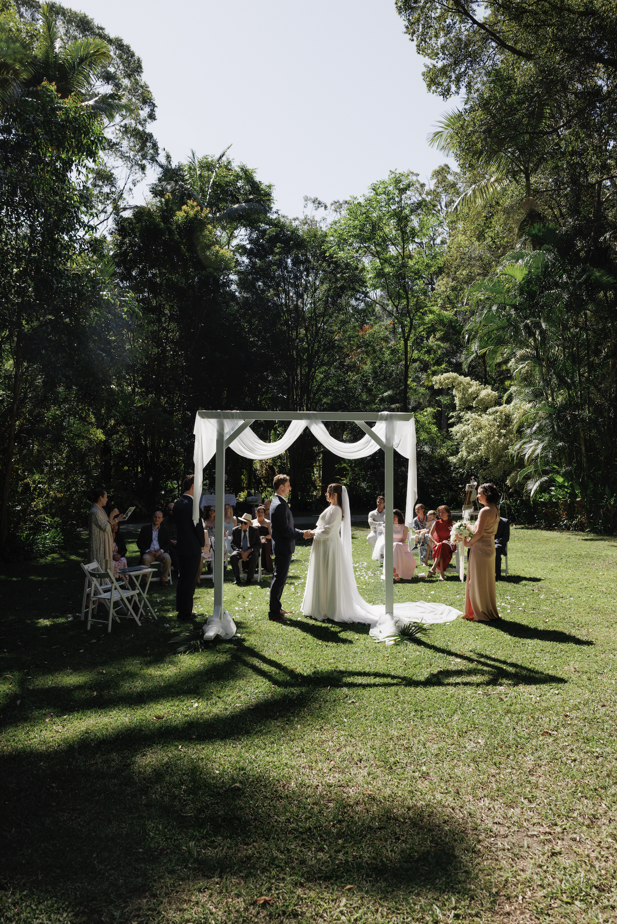 A couple standing and holding hands at their outdoor wedding ceremony, with guests seated behind them in a lush green forest setting.
