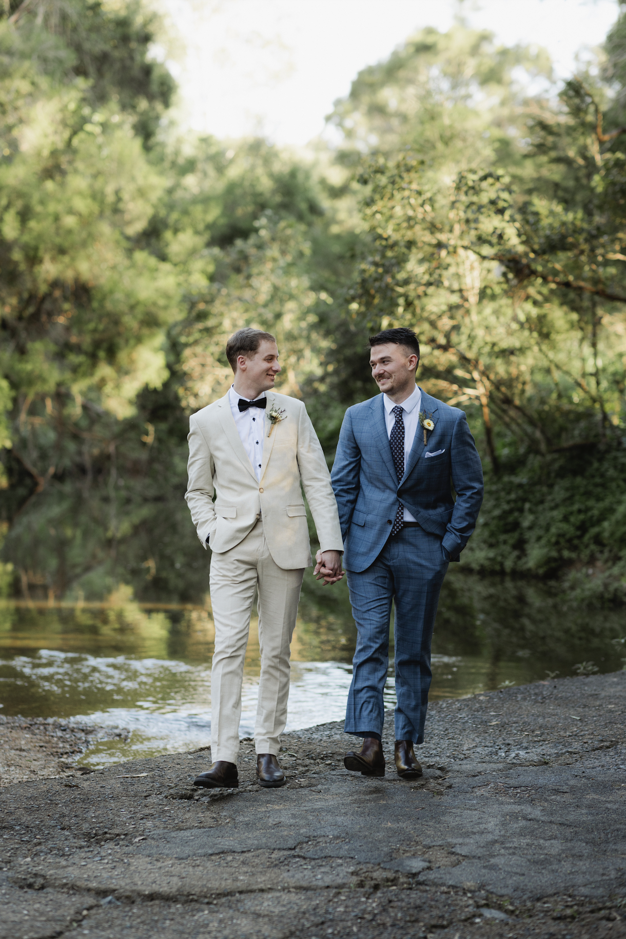 Two men in suits, one in a cream suit and the other in a blue suit, holding hands and walking by a river surrounded by trees.