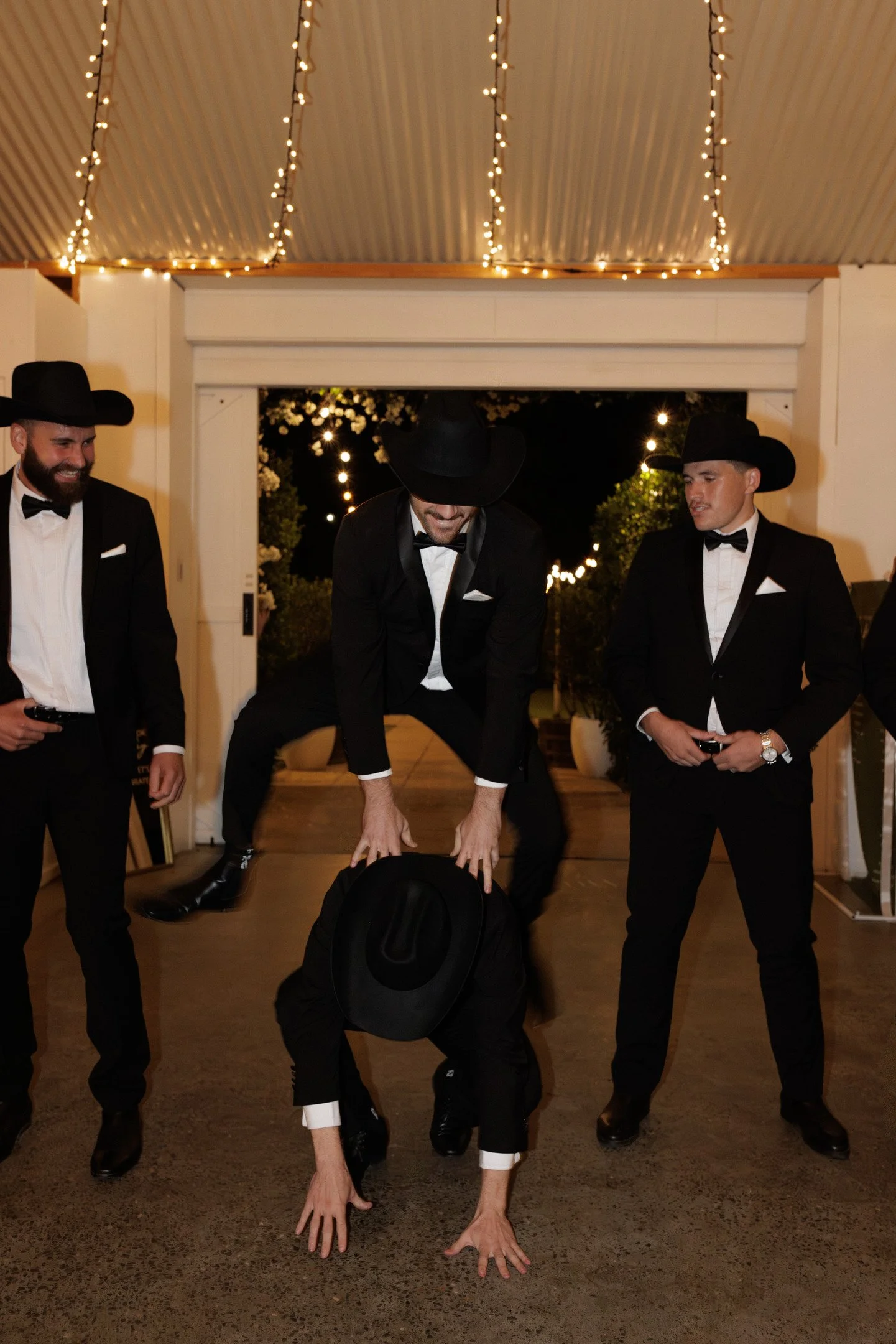 Four men dressed in tuxedos and cowboy hats at a formal event, with one man crouching and touching the ground, and others standing nearby, in a decorated setting with string lights.