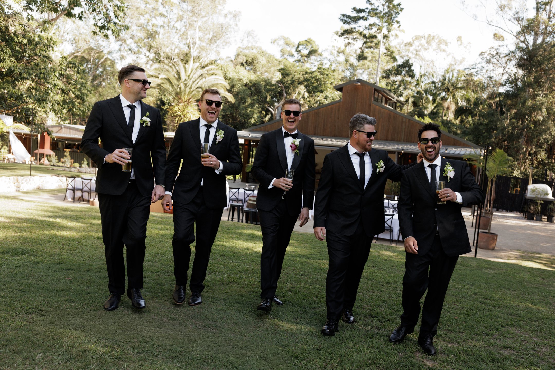 A group of five men in black suits and sunglasses are walking outdoors at a wedding reception, holding drinks and laughing.
