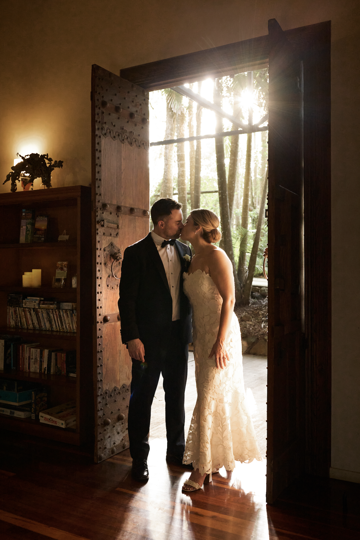 A bride and groom sharing a kiss at the entrance of a rustic venue with trees outside, a wooden door, and a bookshelf inside, illuminated by natural sunlight.