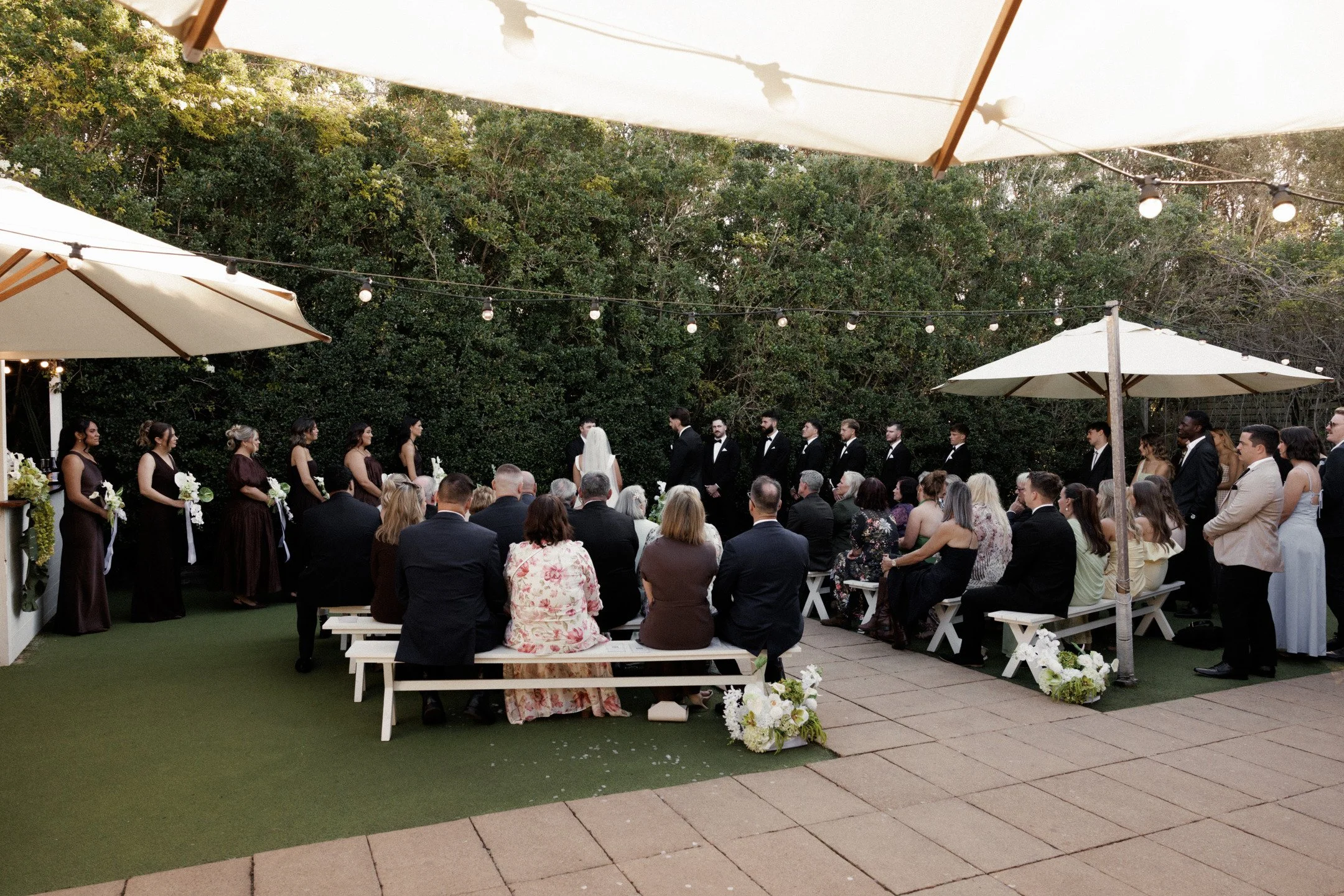 An outdoor wedding ceremony with guests seated and the bride and groom standing at the altar under large white umbrellas, surrounded by string lights and greenery.