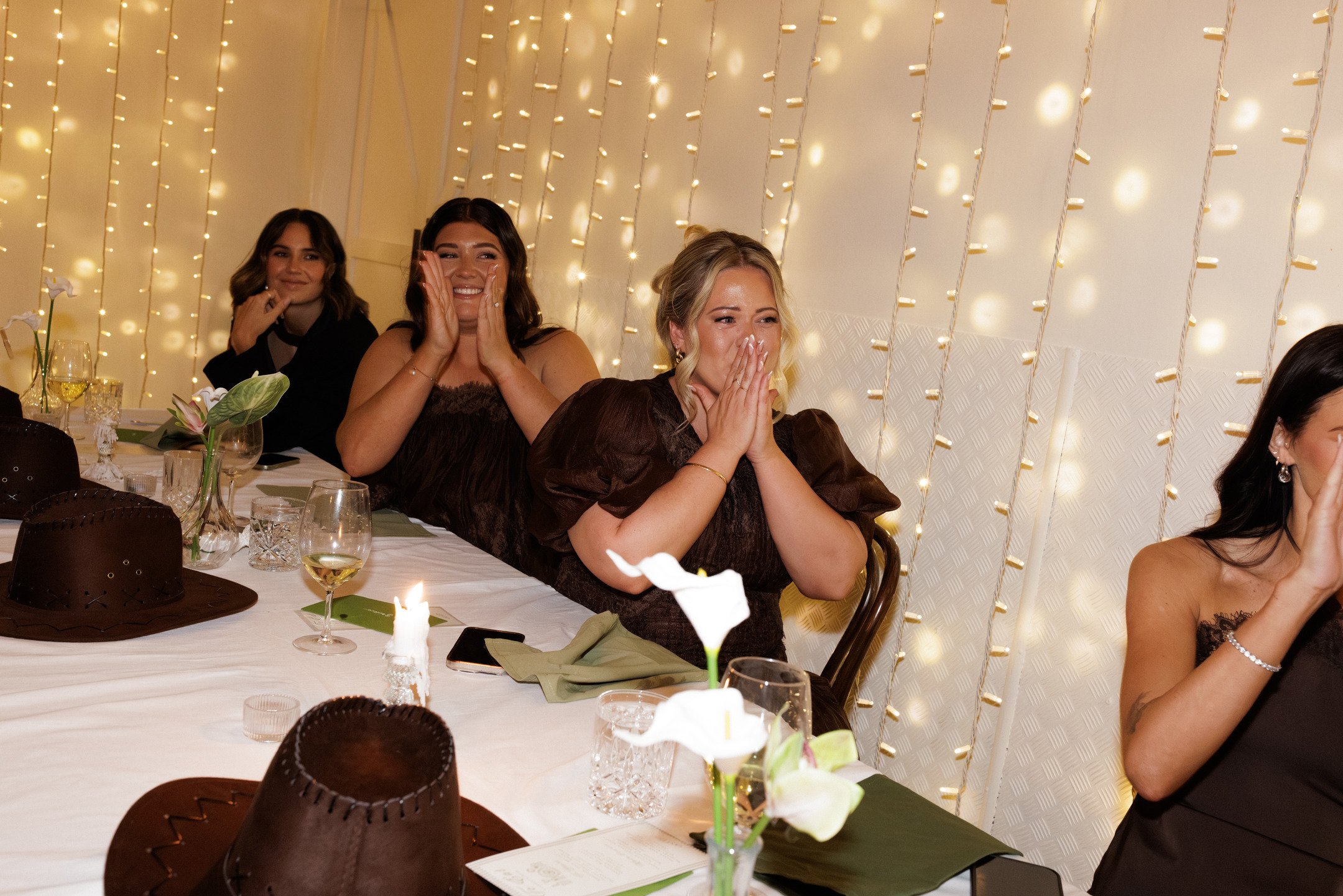 Women seated at a banquet table, reacting with joy and surprise during a celebration, with warm lighting and floral decor.