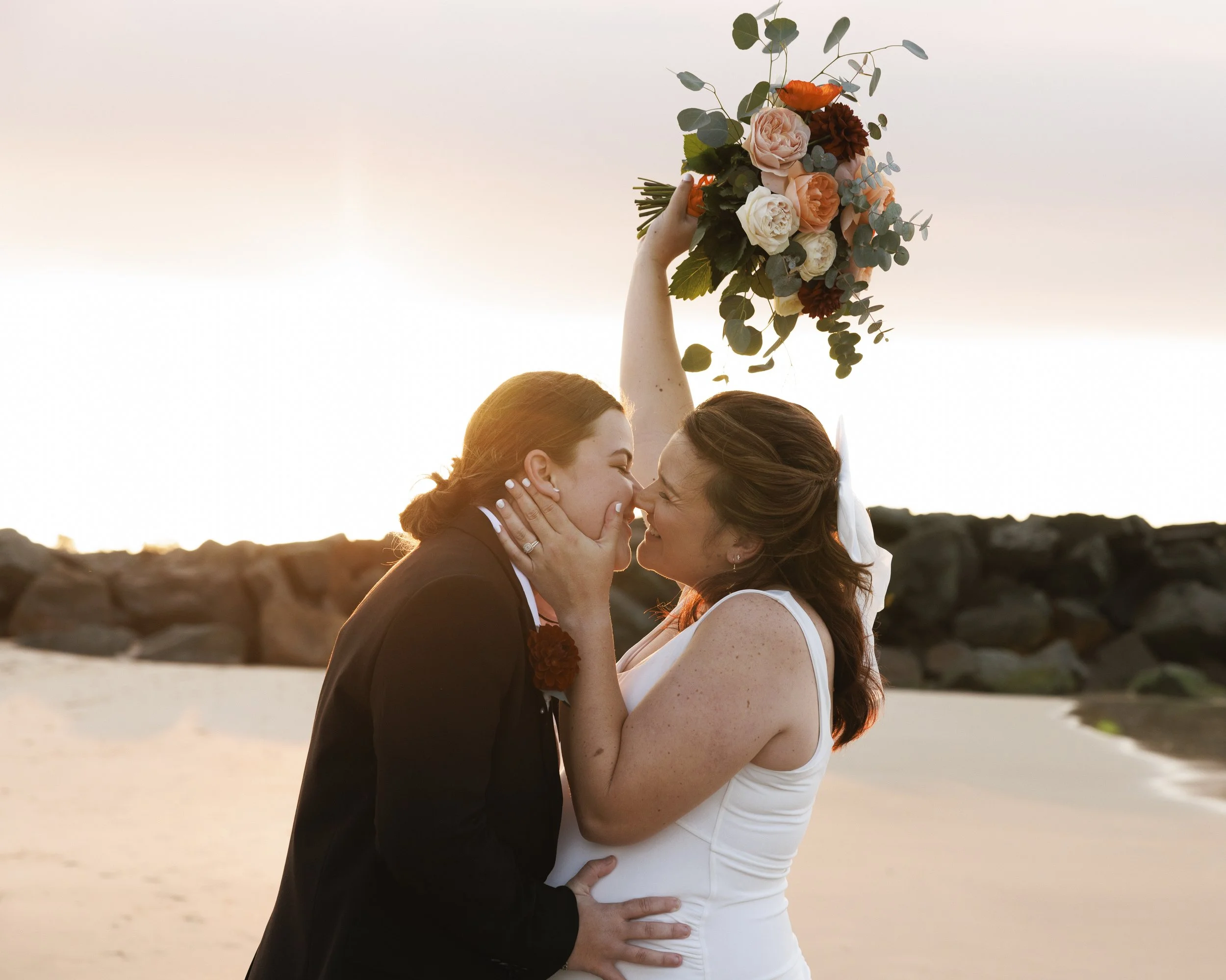 Two women in wedding attire sharing a kiss on the beach at sunset, one holding a bouquet of flowers above them.