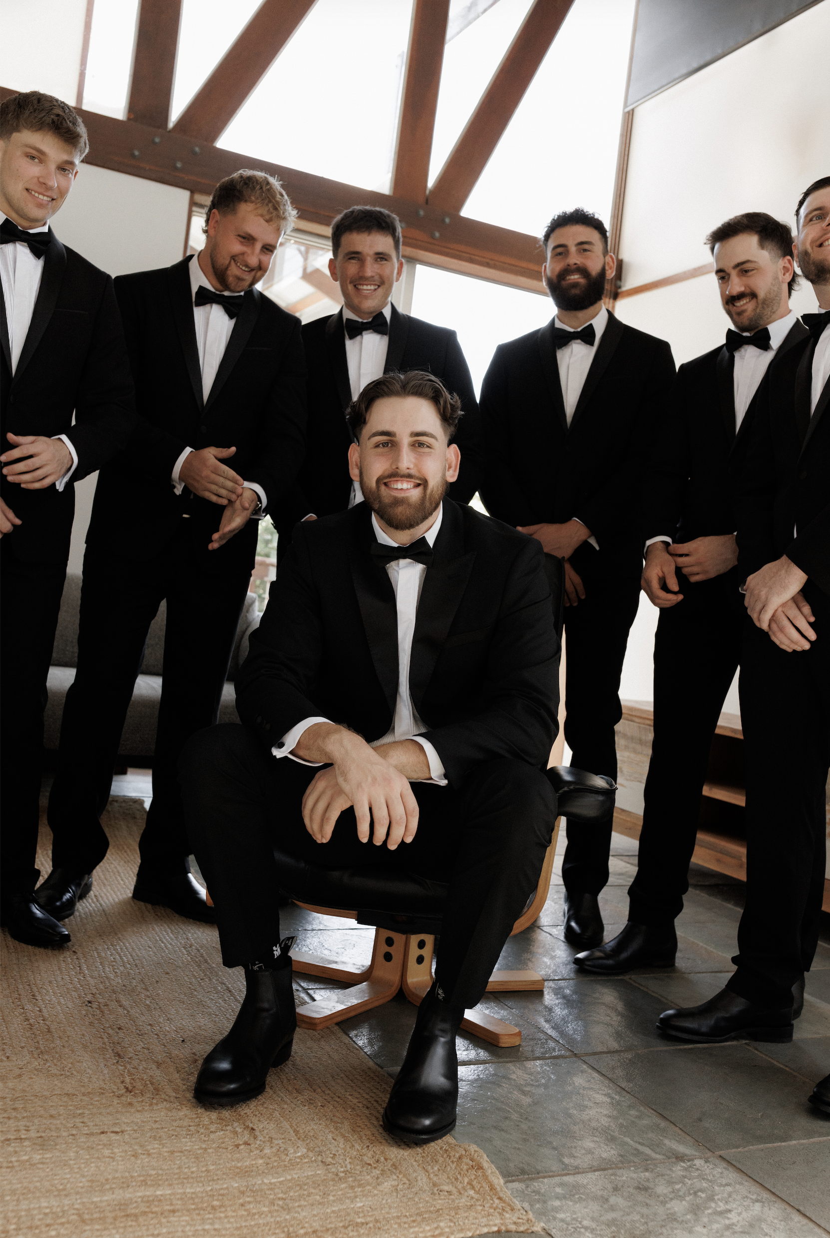 A group of men dressed in black tuxedos and bowties, with one man sitting and smiling in front, inside a modern house with wooden beams and large windows.