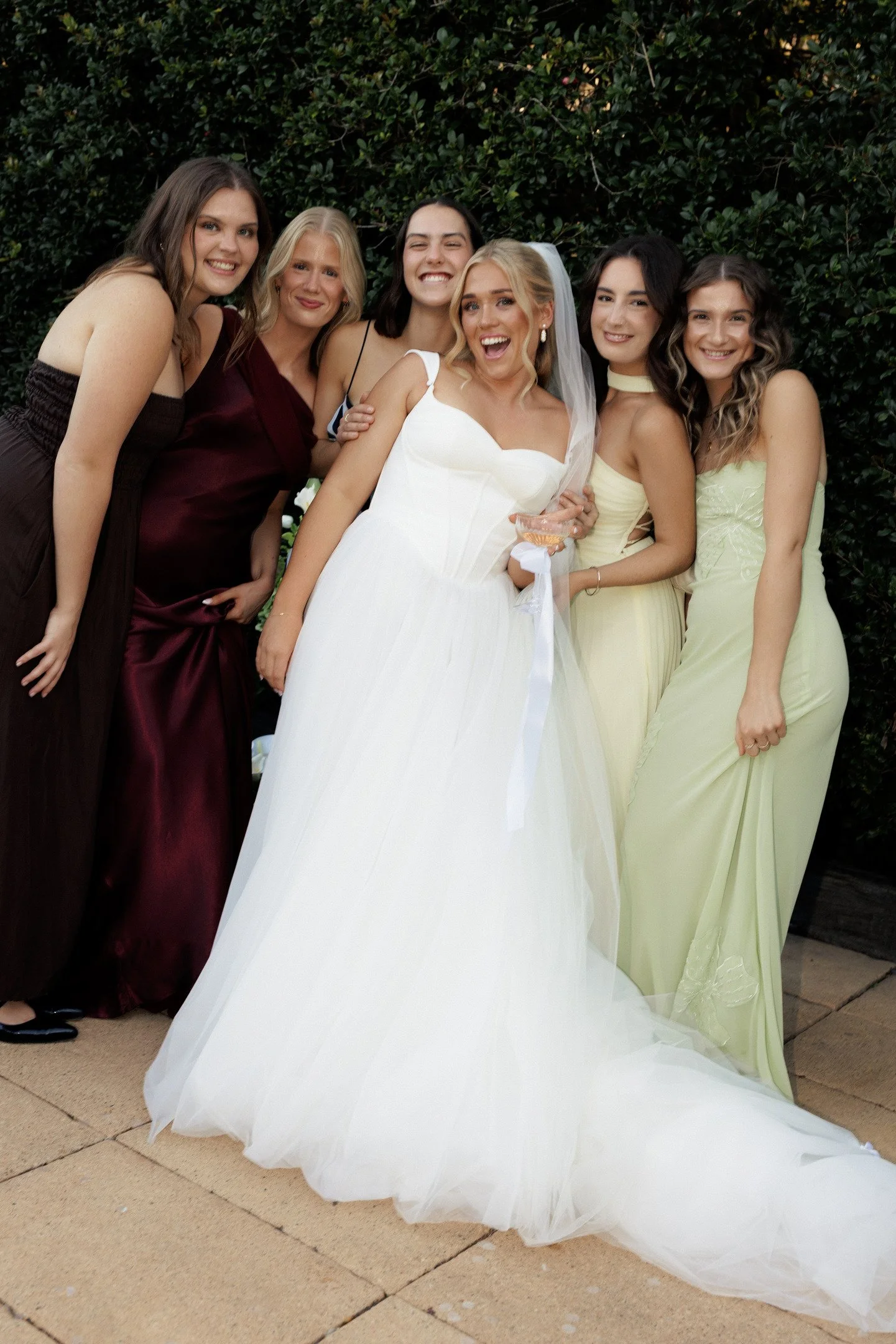 Group of six women, including one bride in a white wedding dress with veil, smiling and posing outdoors against a green leafy background.