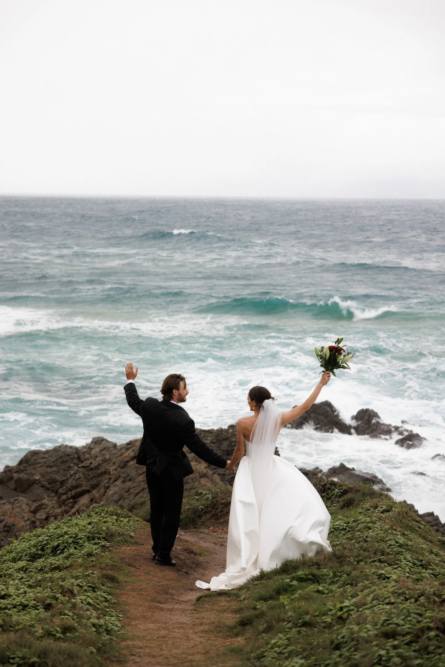 A couple dressed in wedding attire is walking along a rocky coastline with the ocean in the background. The bride is holding a bouquet and raising her arm, while the groom is holding her hand and waving.