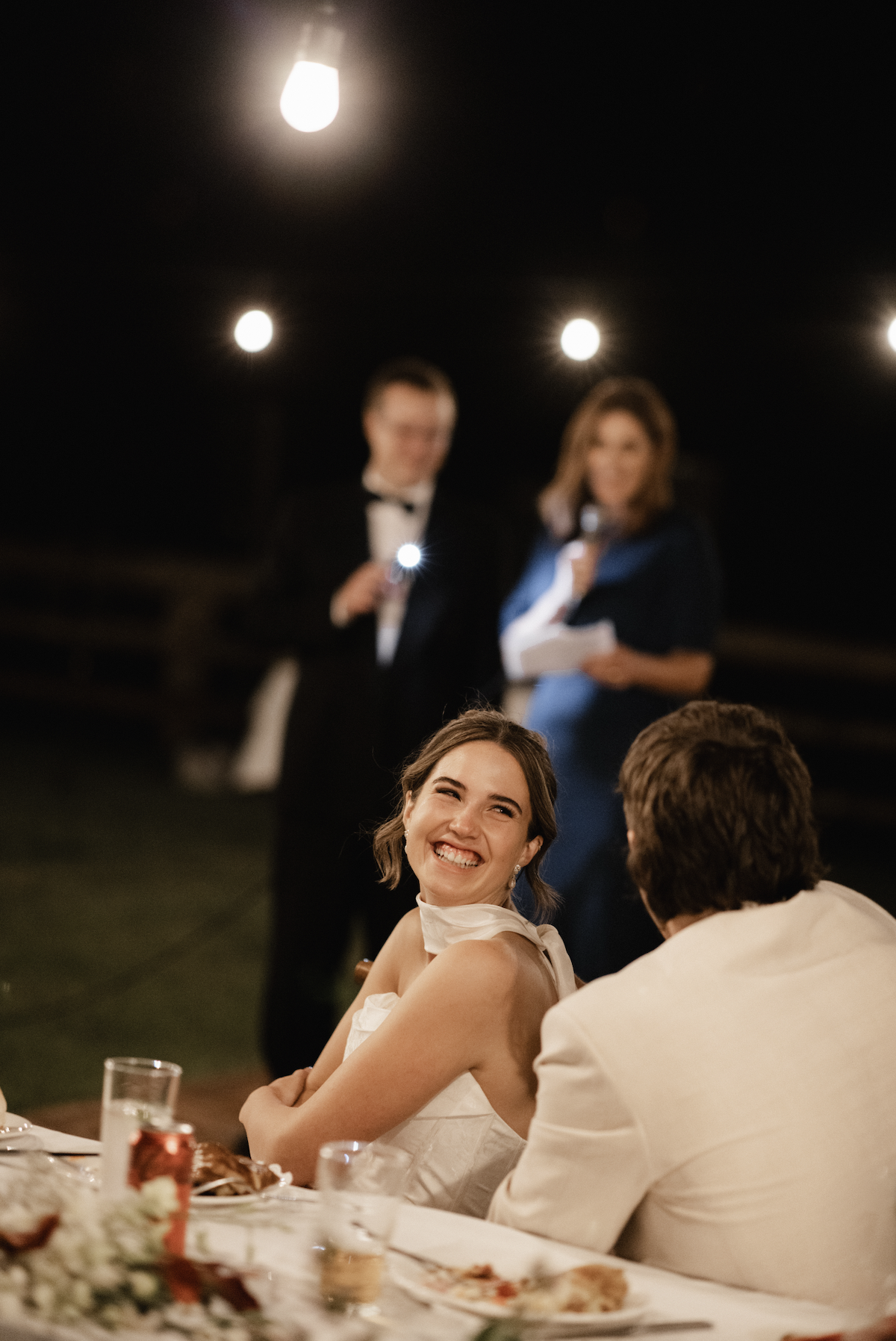 A woman in a white dress smiling and laughing at a dinner table during an evening event, with two blurred people standing and holding papers in the background.