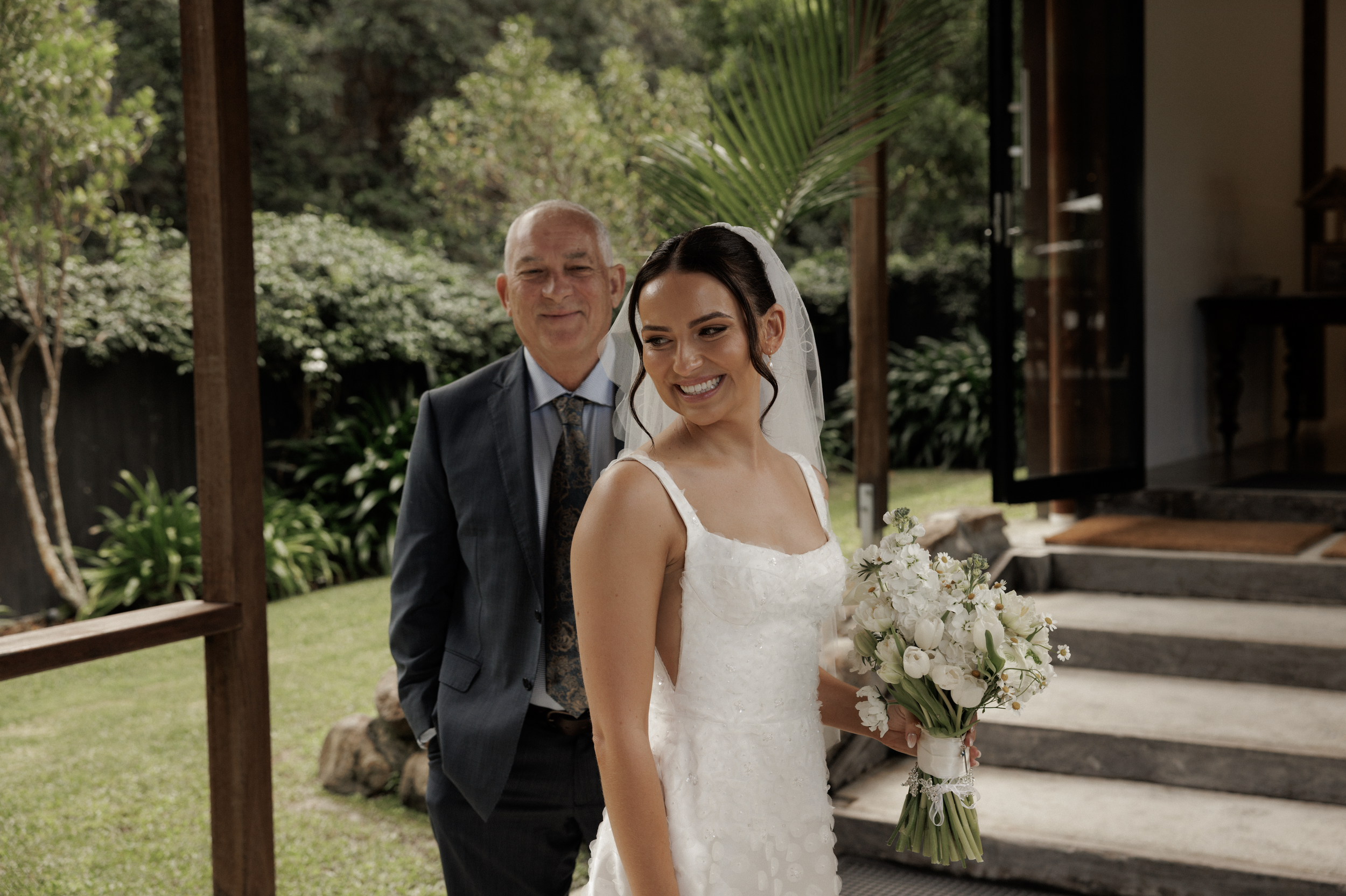 A bride in a white wedding dress holding a bouquet of white flowers, standing outdoors near steps, with a man in a suit smiling in the background, and green foliage and trees surrounding them.