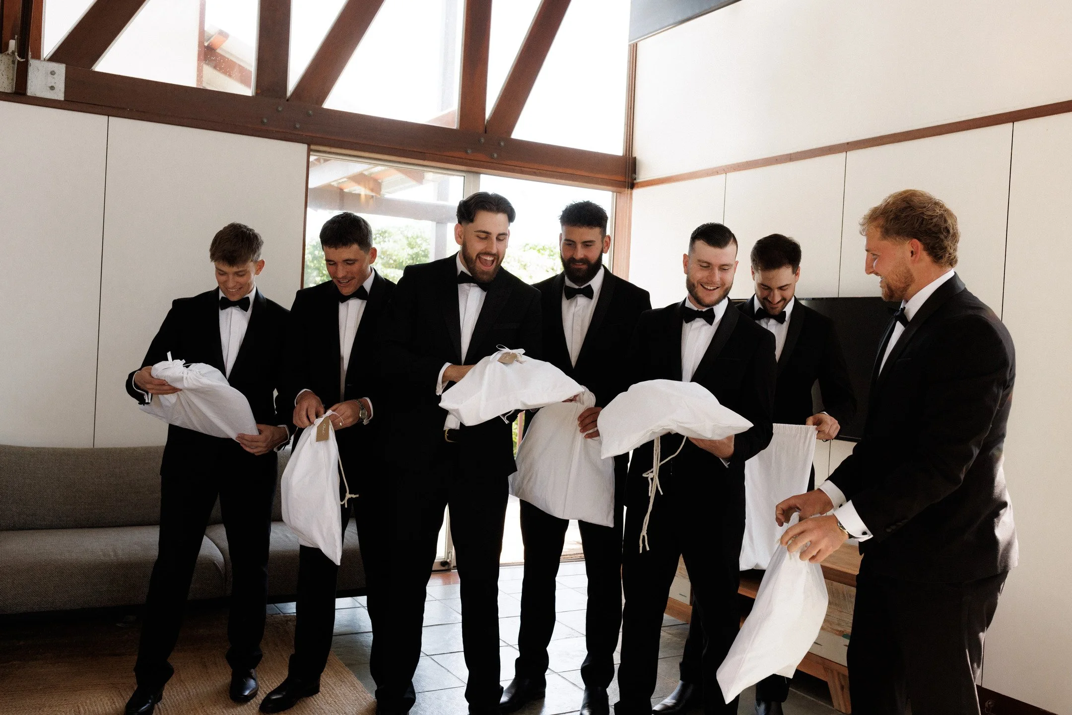 Six men in black tuxedos with bow ties smiling and gathering around white cloth bags in a modern, well-lit room with wooden accents and large windows.
