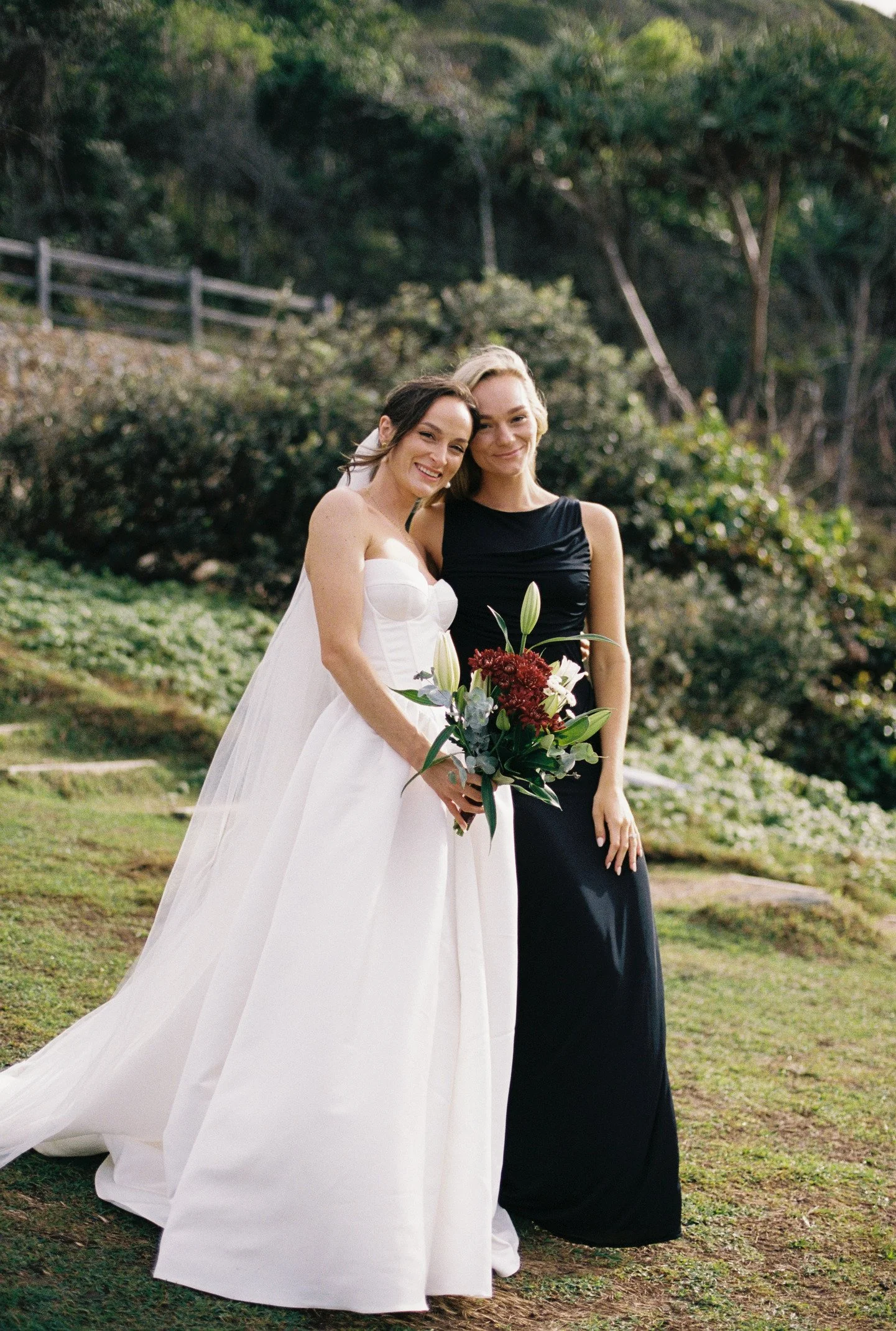 Two women, one in a white wedding dress and the other in a black dress, standing together outdoors in a garden with greenery and trees in the background. The woman in the wedding dress is holding a bouquet of flowers and smiling, while the other woma