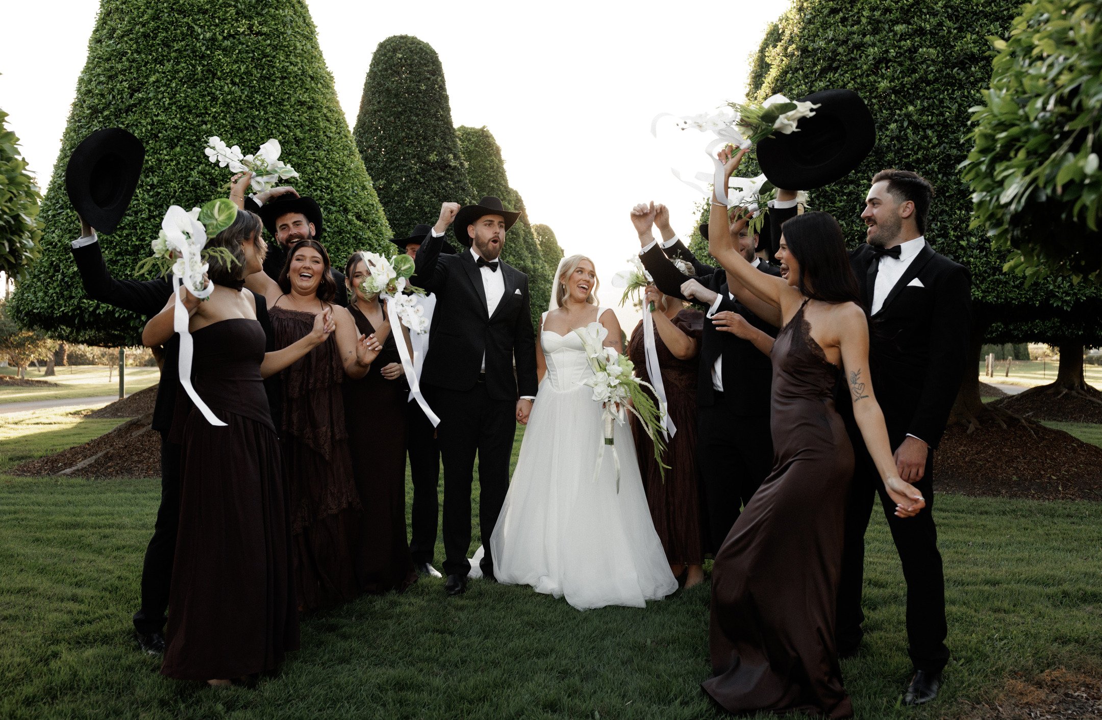 A group of wedding guests in formal attire celebrating outdoors with a bride and groom in the center, surrounded by tall, trimmed bushes and trees.