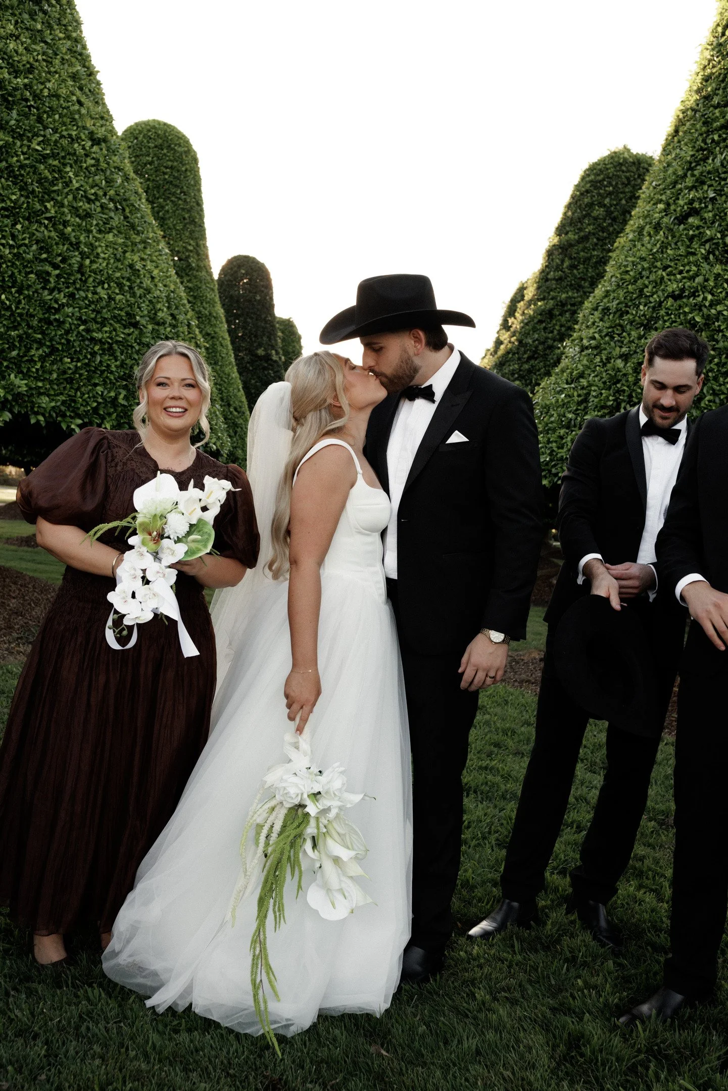 A bride and groom share a kiss during their wedding ceremony outdoors, with bridesmaids and groomsmen standing nearby, surrounded by neatly trimmed bushes.