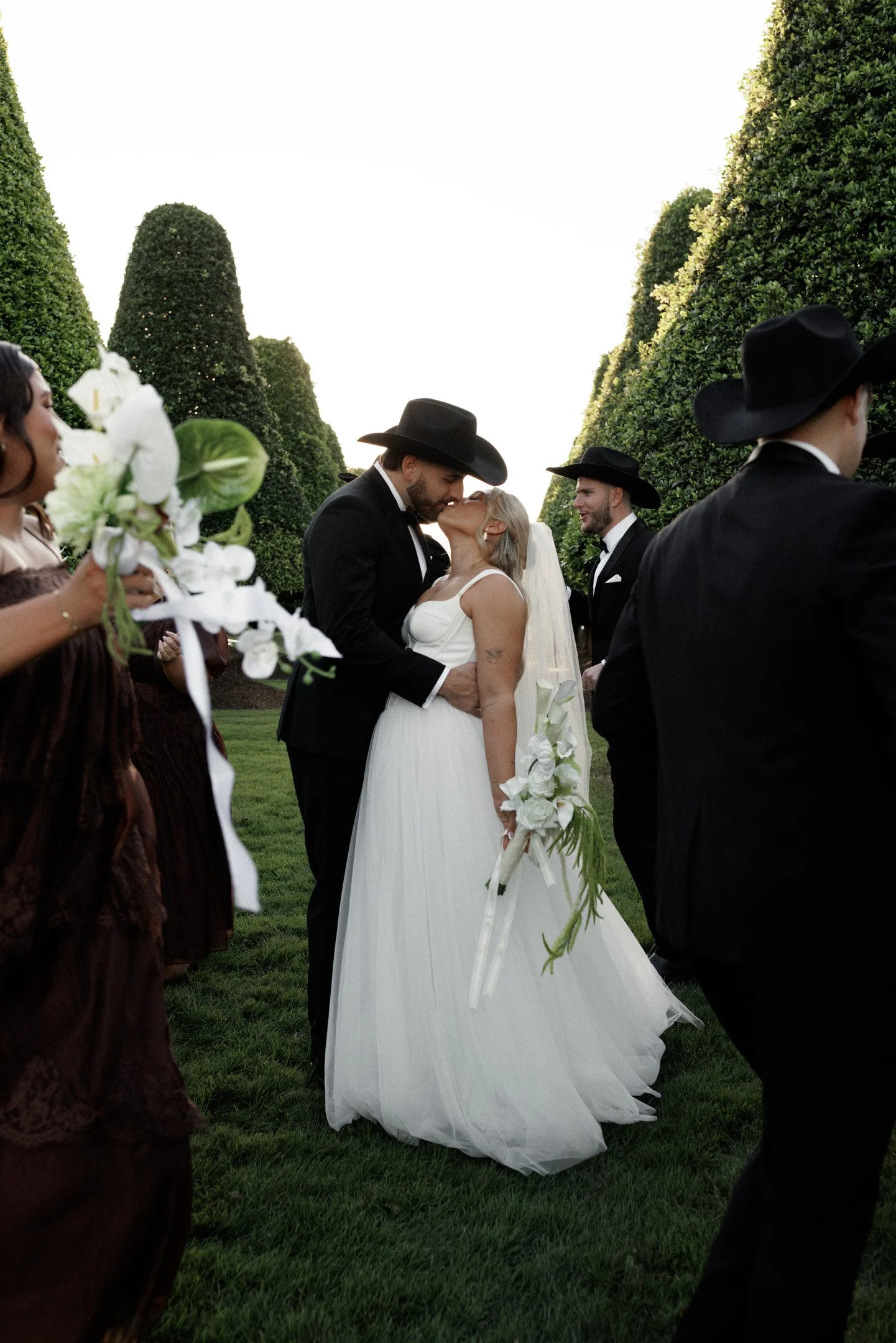 A bride and groom sharing a kiss during their outdoor wedding ceremony, surrounded by bridesmaids and groomsmen, with tall green hedges and trees in the background.