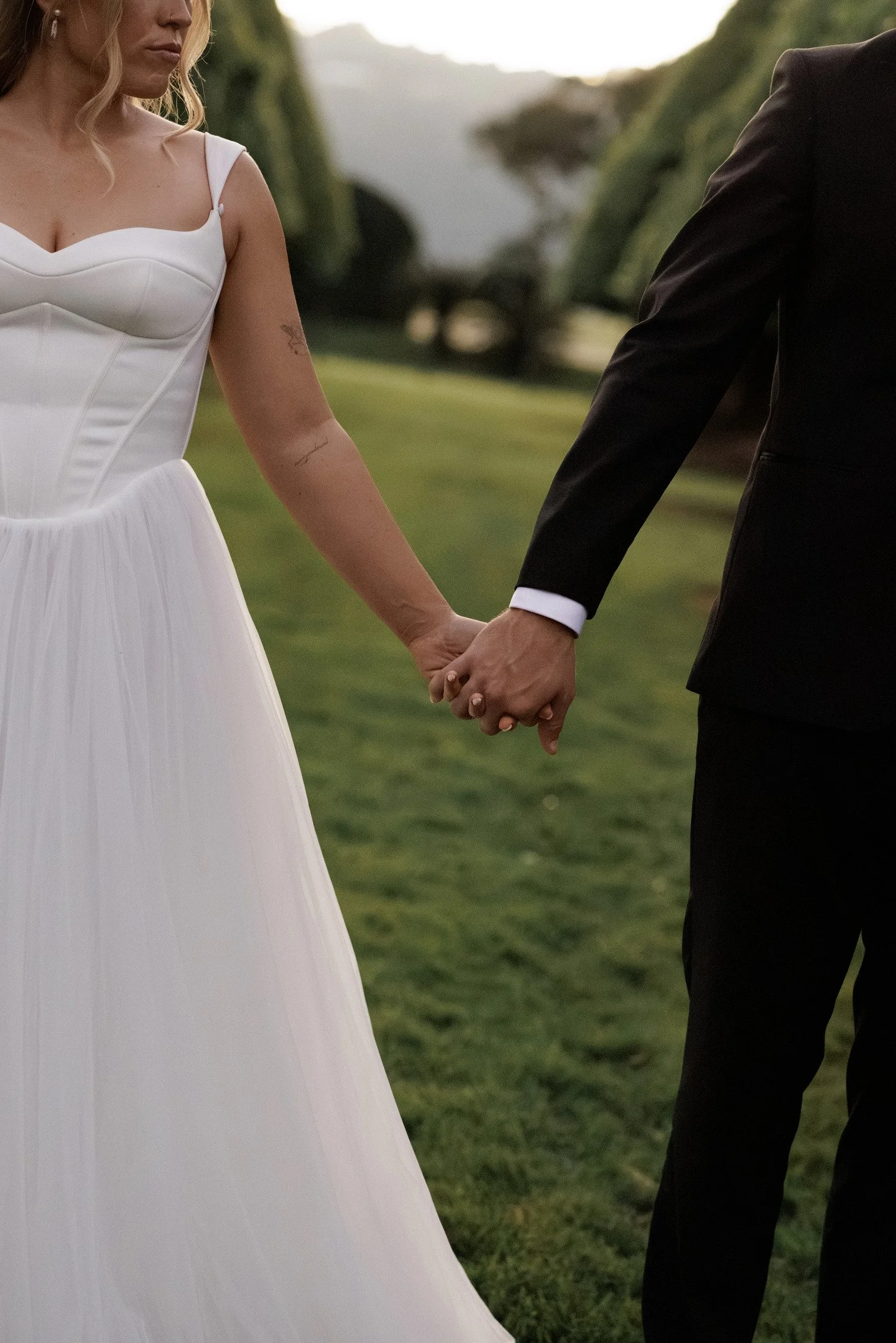 A bride and groom are holding hands outdoors, with a grassy area and trees in the background. The bride is wearing a white wedding dress and the groom is dressed in a black suit.