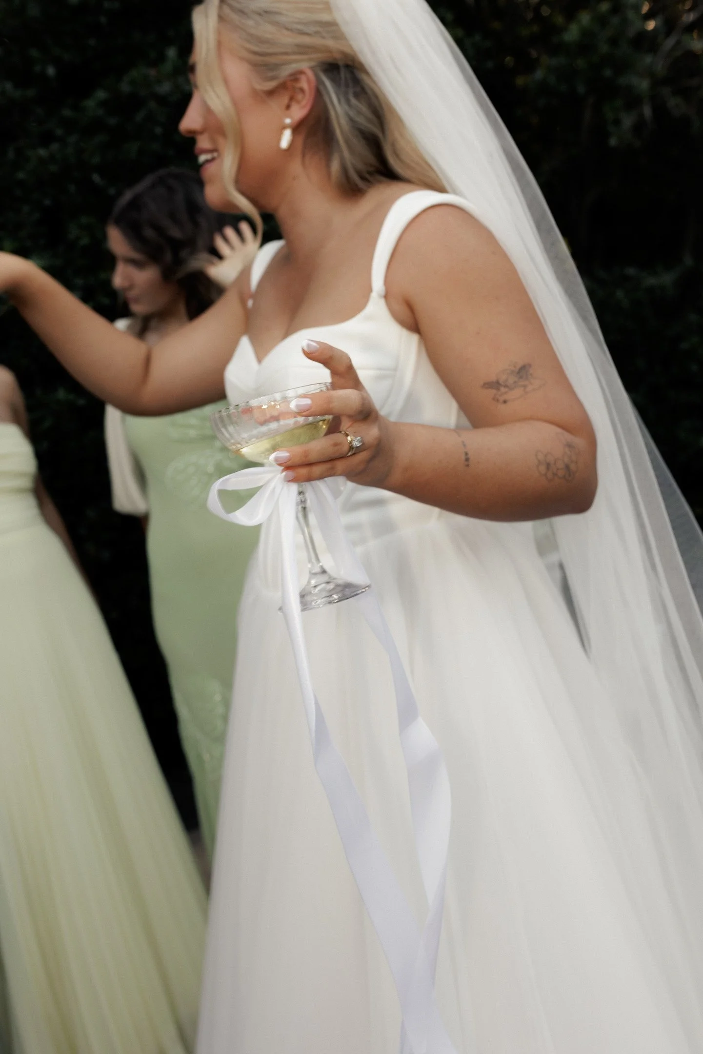 A woman in a white wedding dress with tattoos on her arms holding a glass of champagne, smiling, with a bride and groom in the background outdoors.
