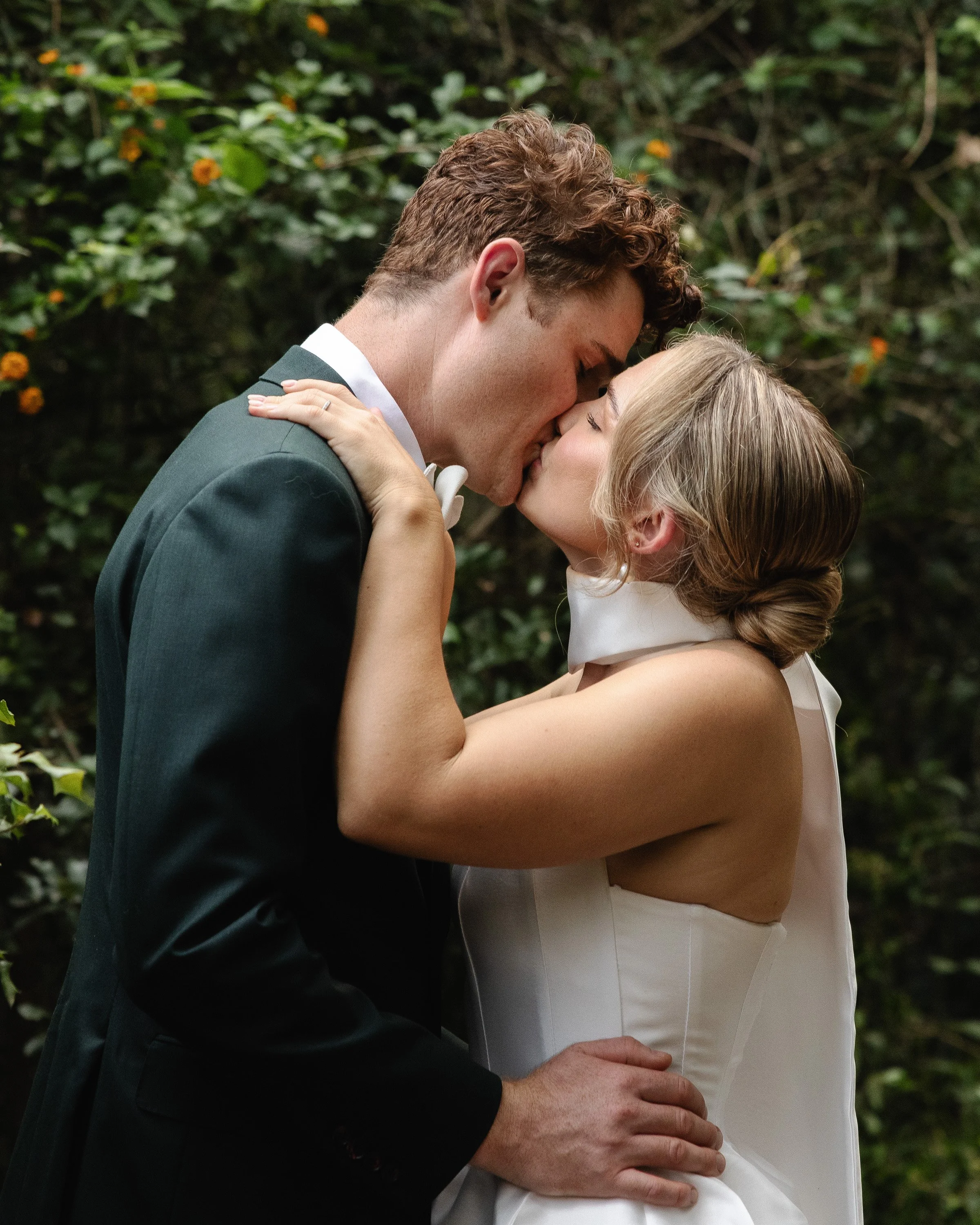 A couple kissing outdoors, with the man wearing a dark suit and the woman in a white dress, surrounded by greenery - Elevated Weddings