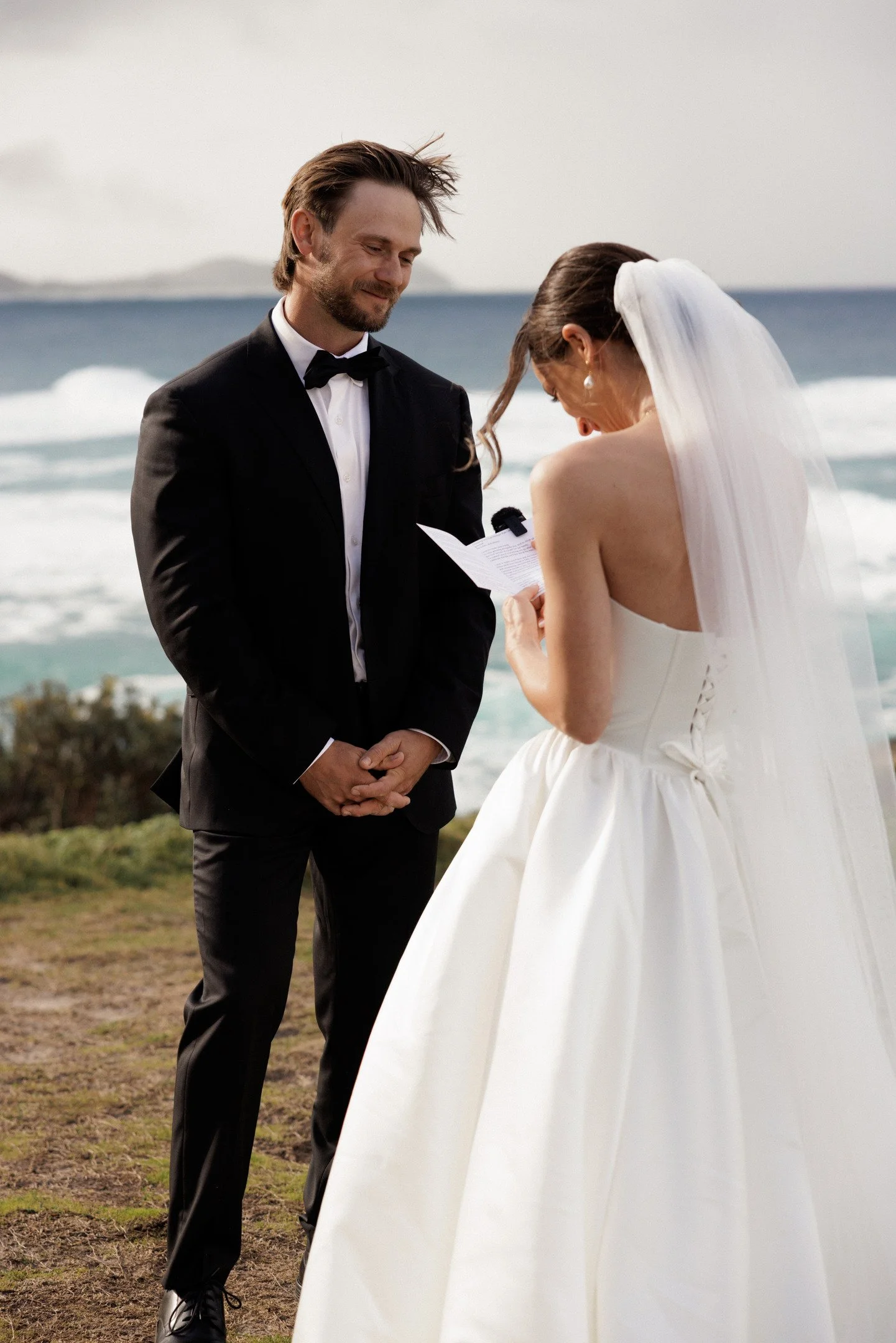 A bride and groom exchange vows on a beach during their wedding ceremony, with the ocean and sky in the background.