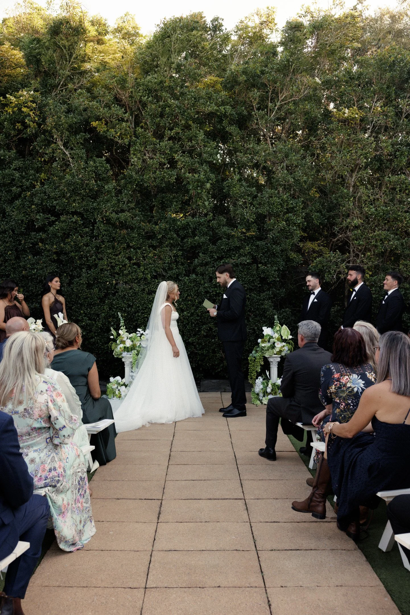 A wedding ceremony outdoors with a bride and groom exchanging vows, surrounded by guests seated on each side, with a lush green tree backdrop.