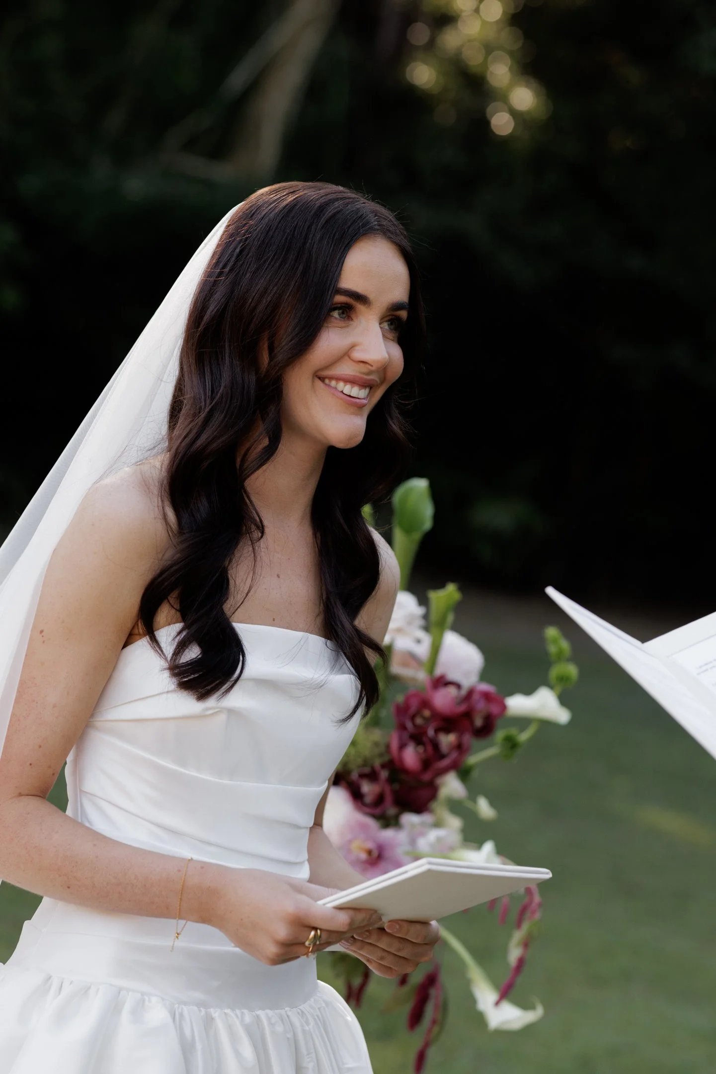 A smiling woman with dark wavy hair, wearing a white strapless dress and a white veil, holding a small booklet or card, standing outdoors with flowers and greenery in the background.